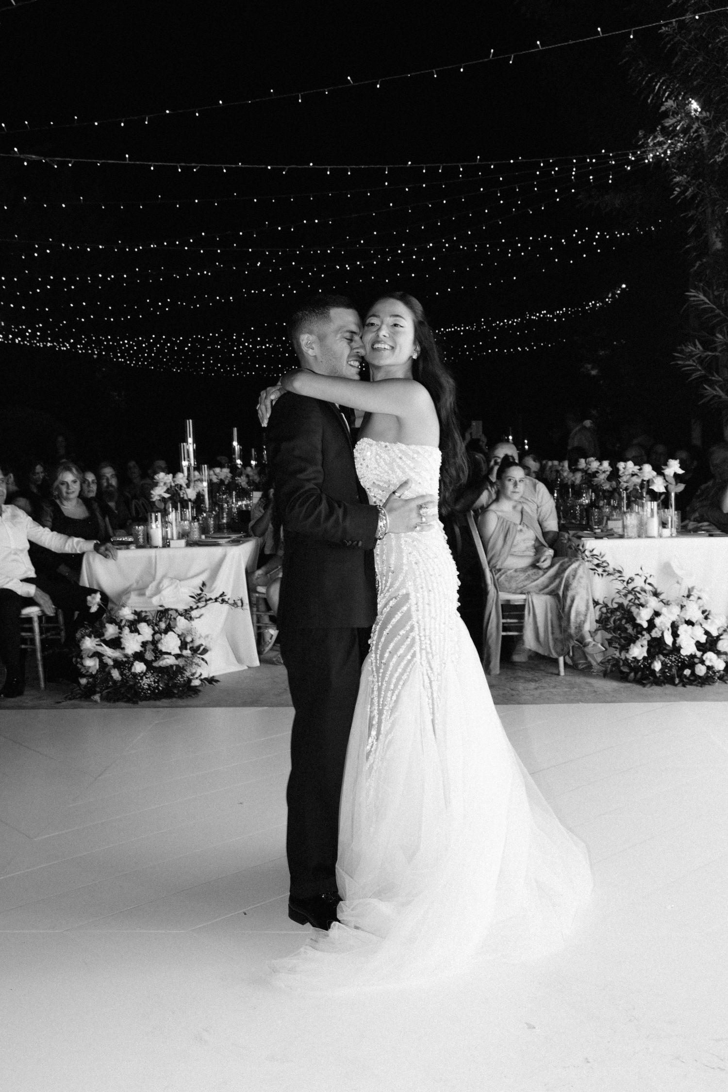 Black and white photo of a bride and groom sharing their first dance under string lights at an outdoor wedding reception.