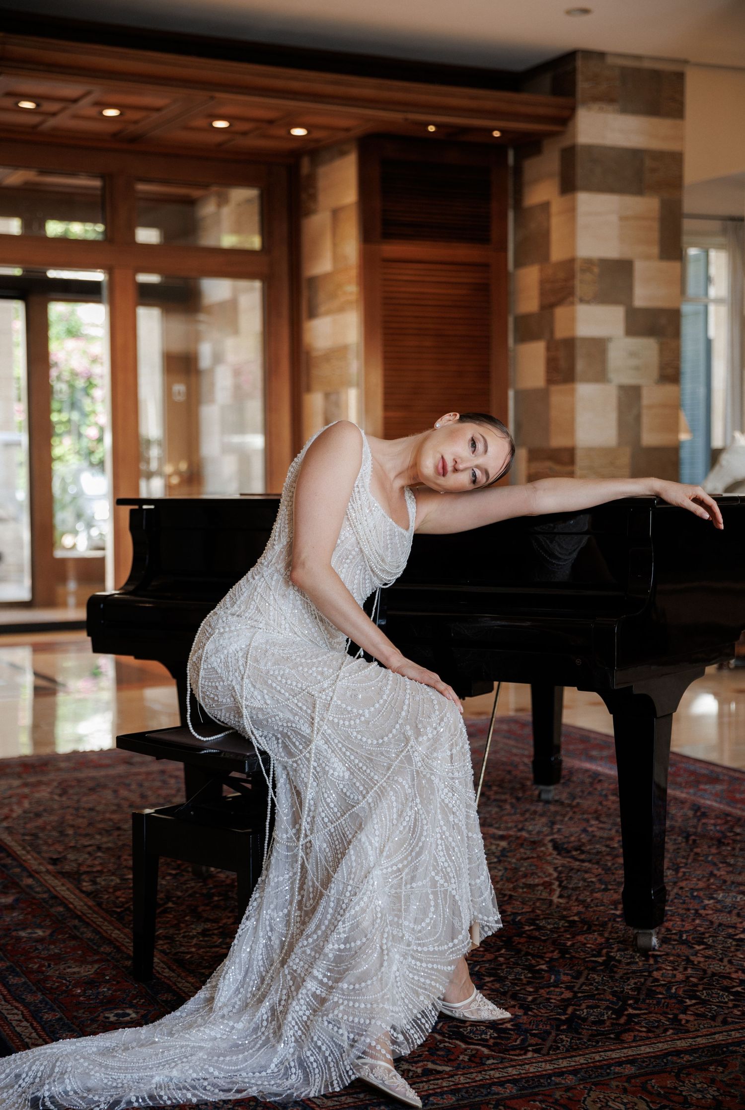 Bride in a beaded white gown leans gracefully on a grand piano in an elegant venue.