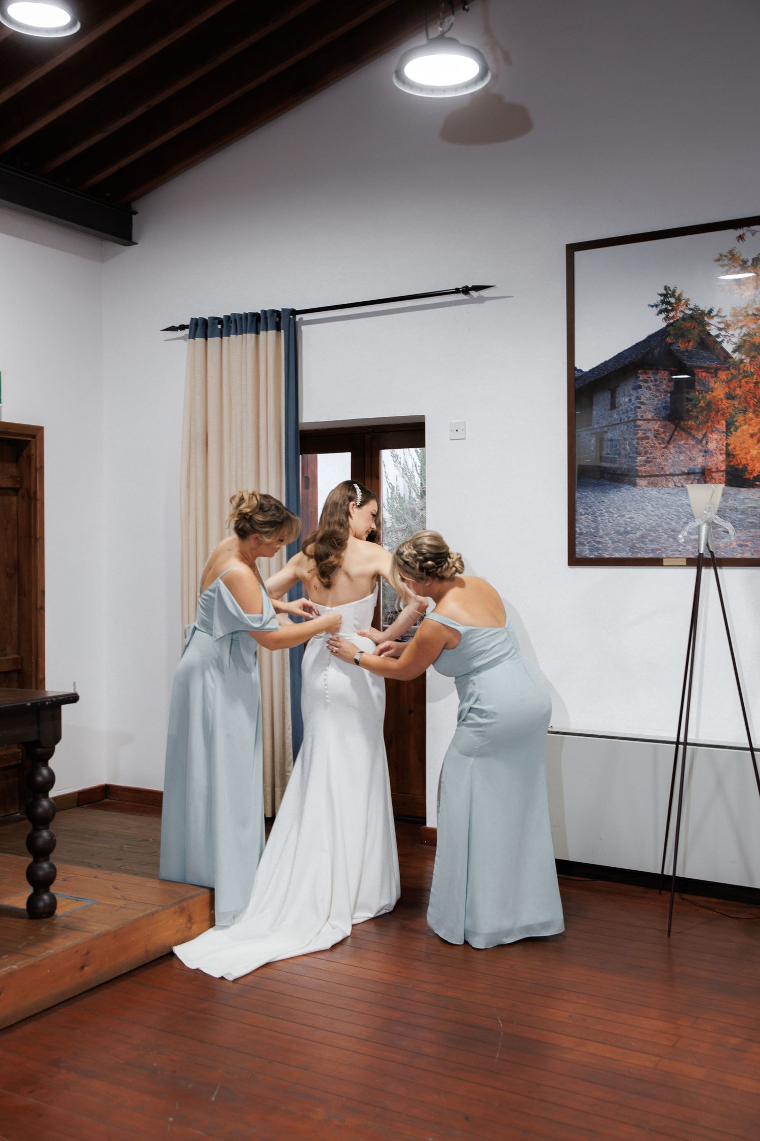 Bridesmaids in light blue dresses help with final adjustments to a white wedding gown in a room with hardwood floors.