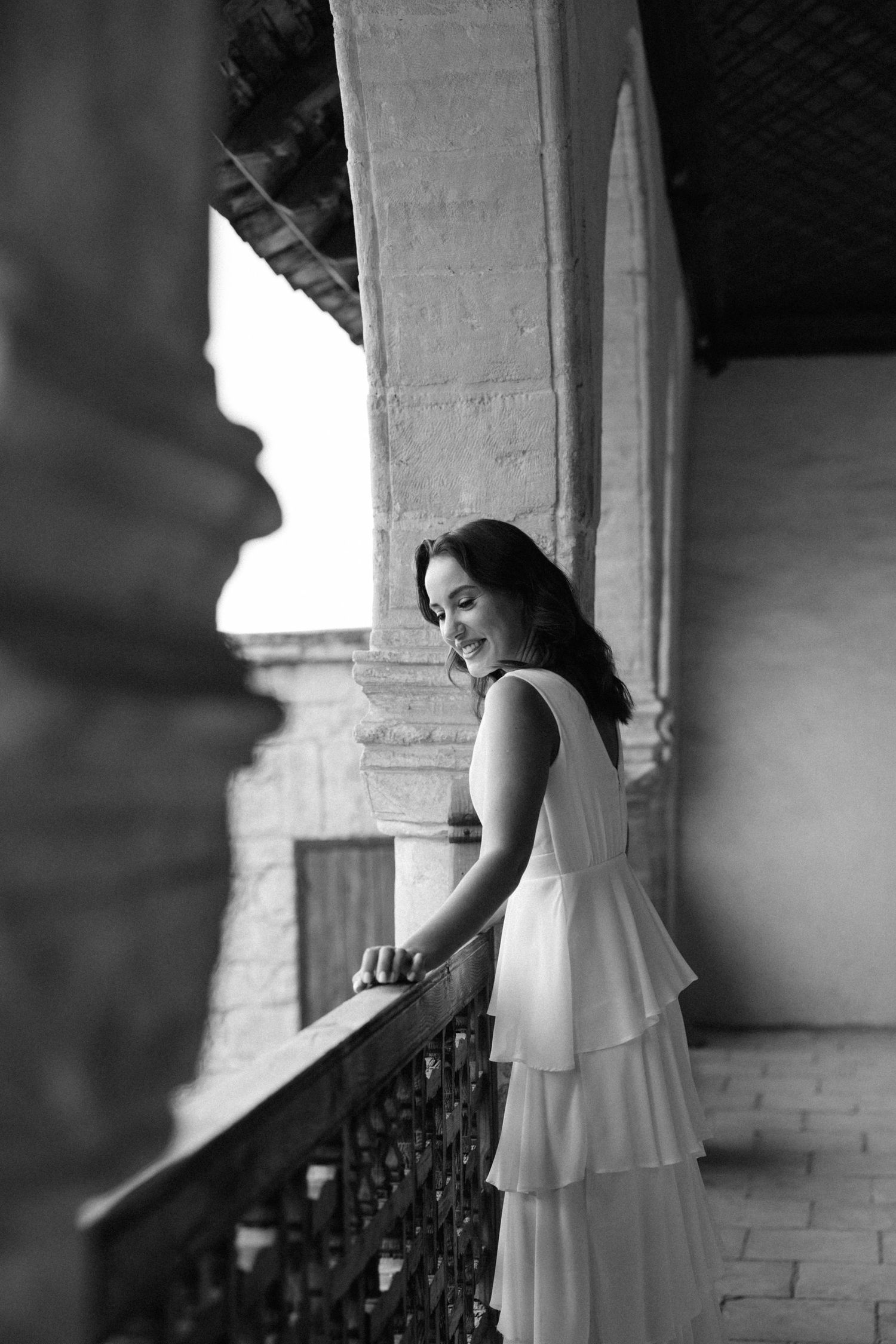 Black and white artistic photograph showing a dancer in a flowing white dress moving gracefully by an ornate railing.