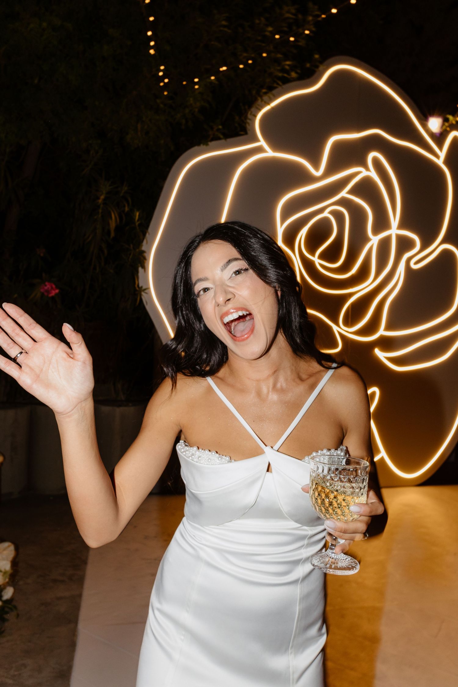 Bride in white dress holding champagne glass, laughing in front of neon rose light decoration at wedding reception.