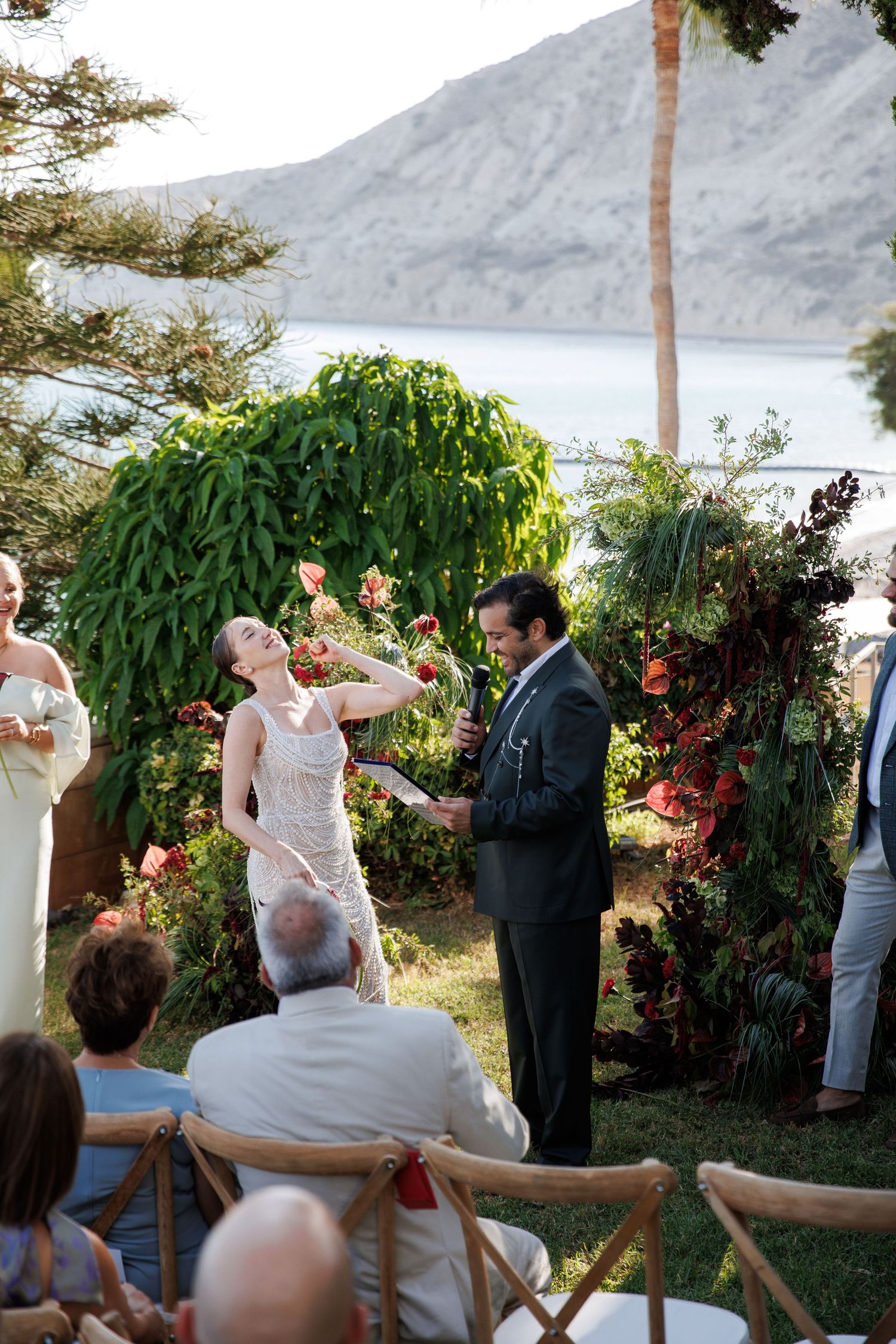 Joyful bride laughs during outdoor wedding ceremony with lush tropical floral arch and mountain lake backdrop.