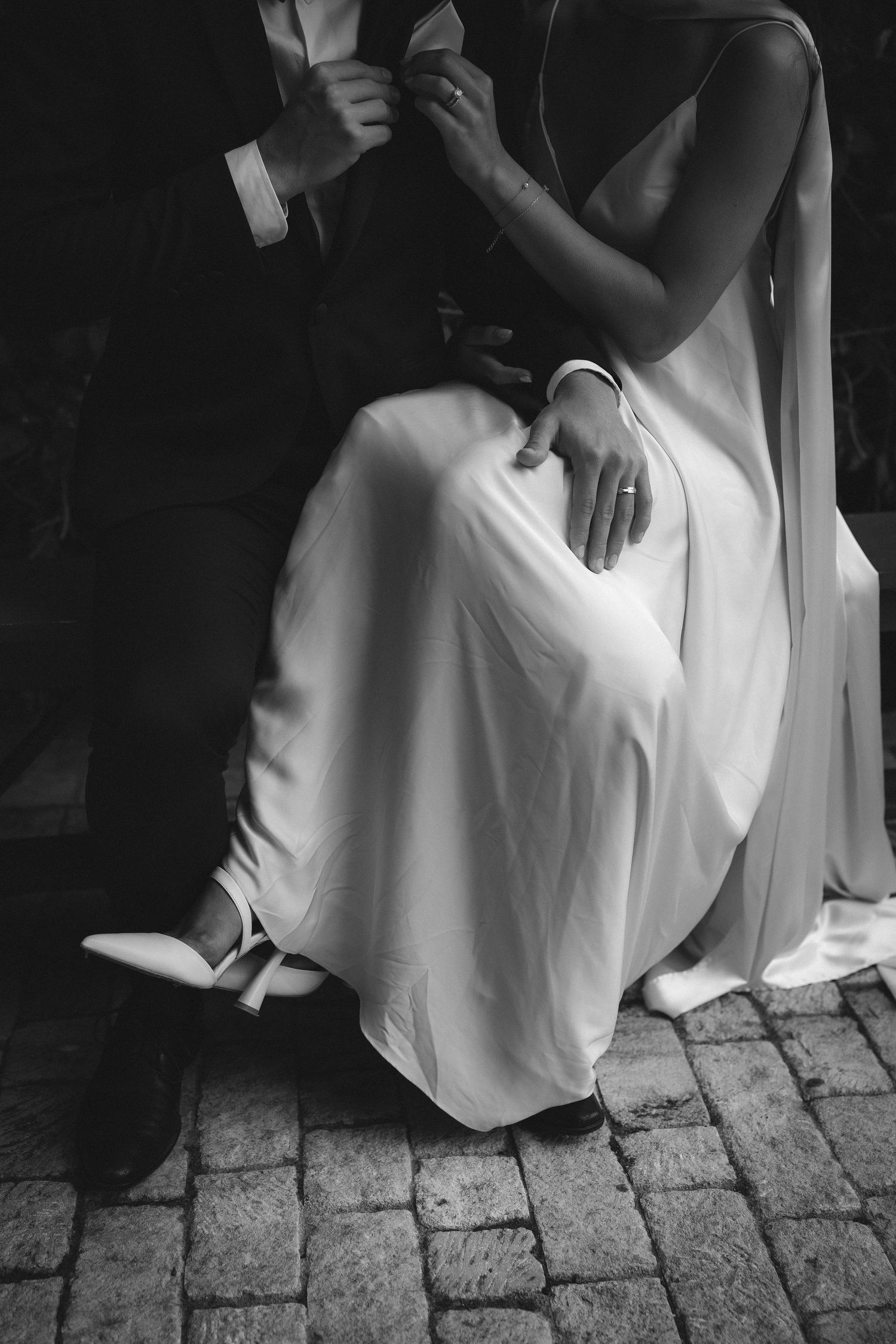 Black and white photo of someone in a flowing white dress and heels sitting on cobblestone pavement.