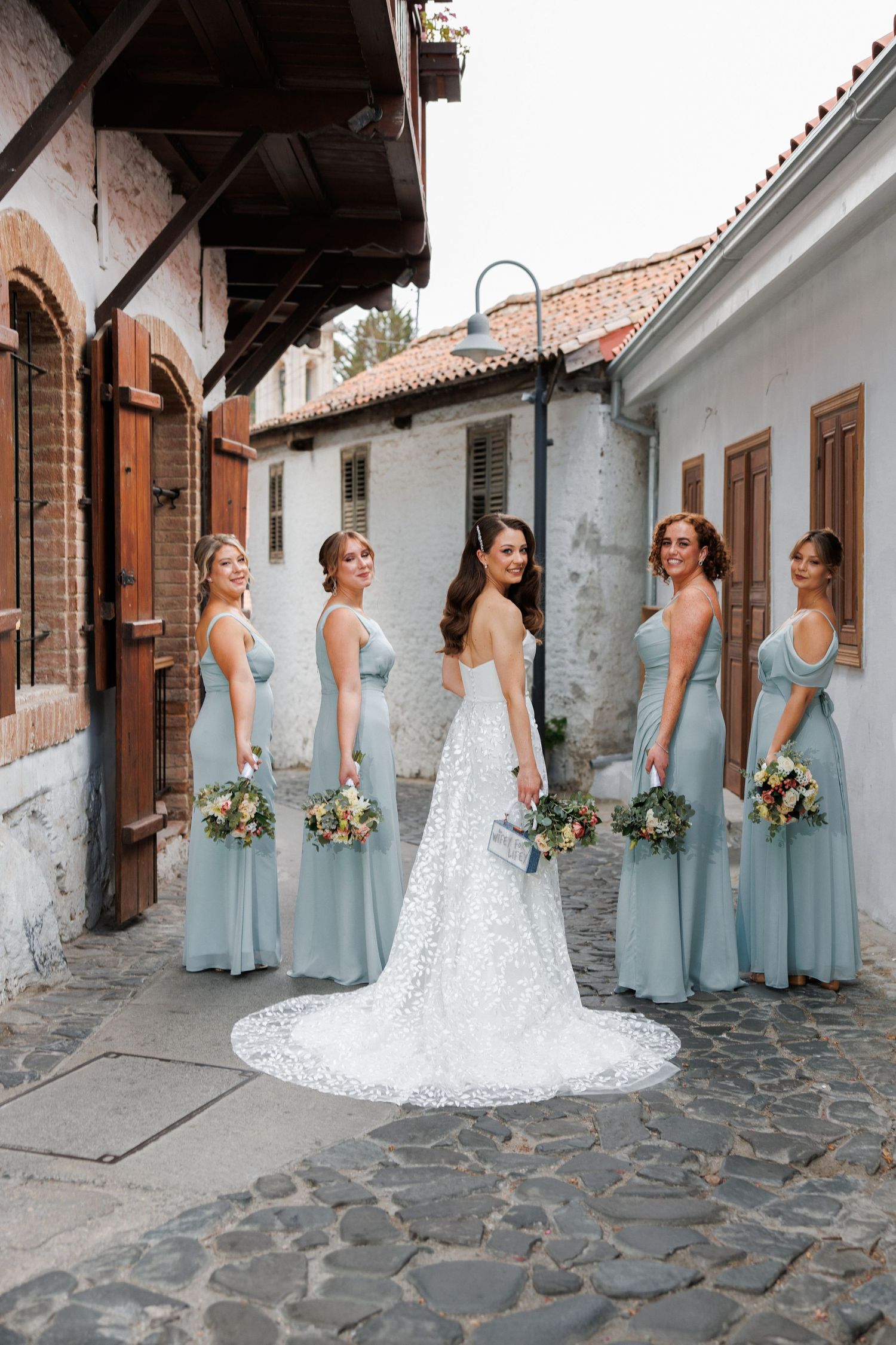 Wedding party poses in light blue bridesmaid dresses with bouquets against rustic stone wall backdrop.