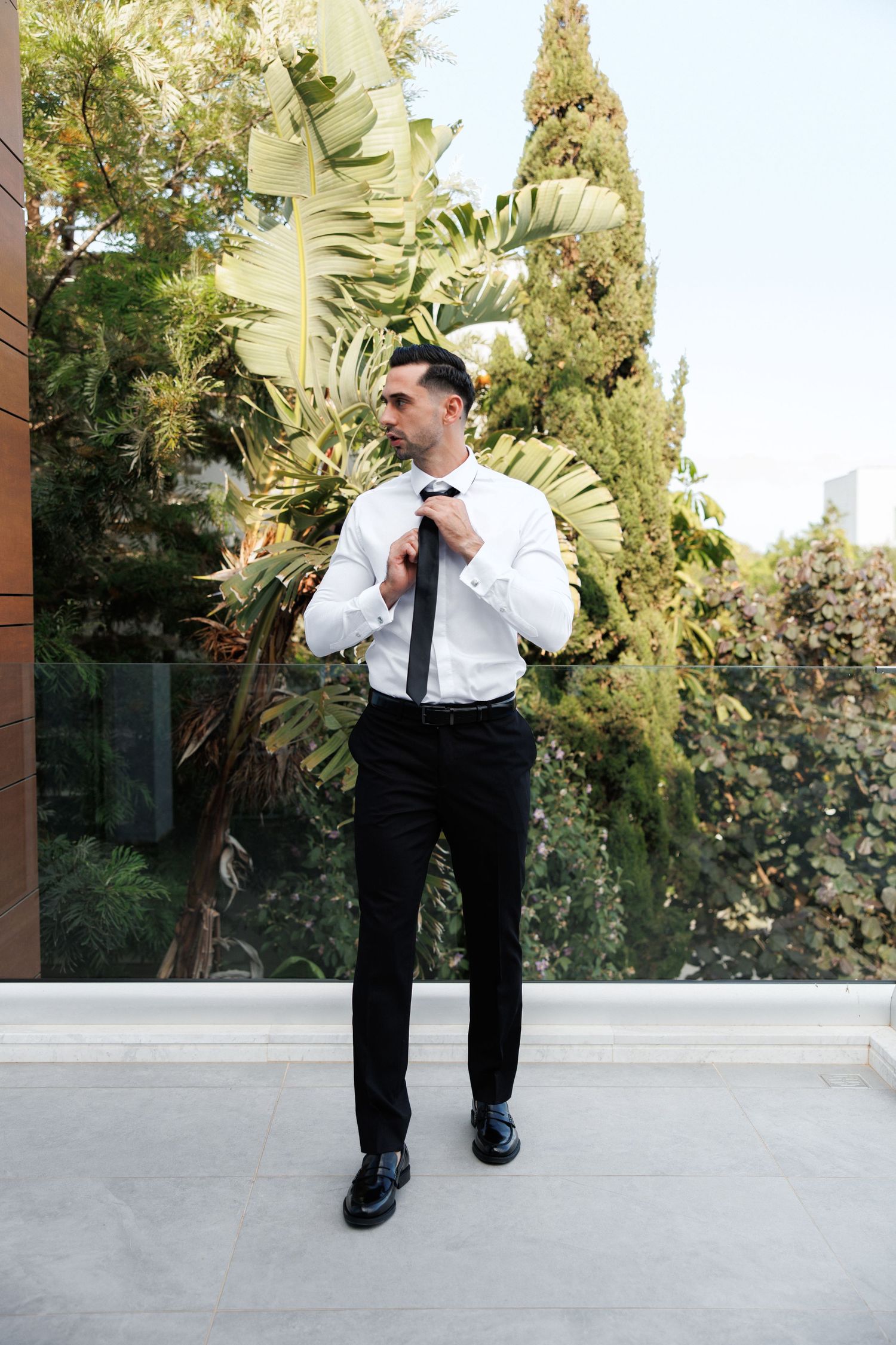 Formal portrait of someone adjusting their tie while standing outdoors near tropical plants and cypress trees.