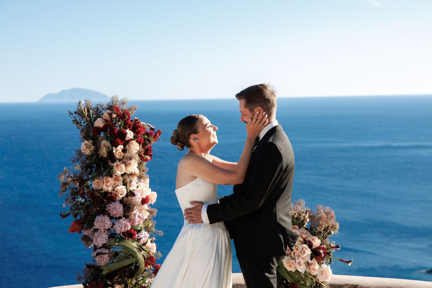 Bride and groom embrace at seaside wedding altar adorned with lush floral arrangement overlooking blue ocean.