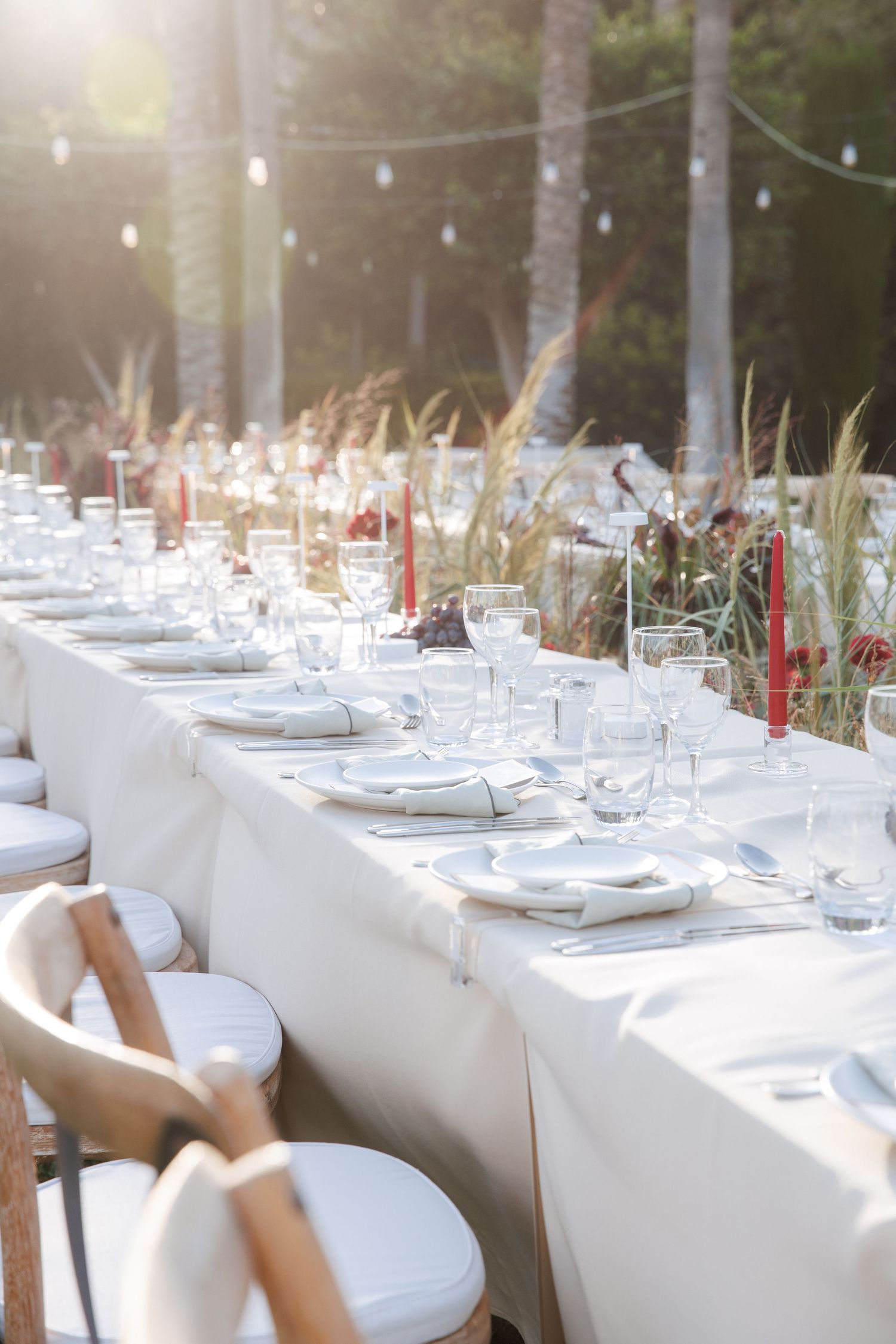 Elegant outdoor dining table set with white linens, glassware, and red candles surrounded by lush greenery.