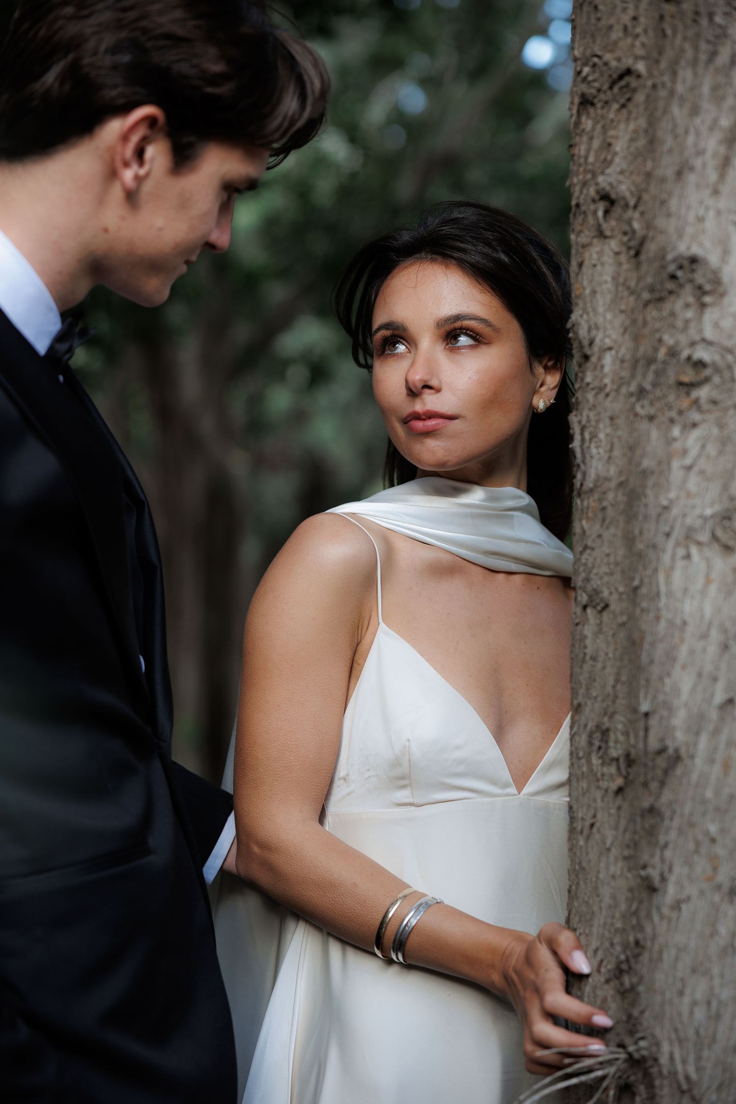 Romantic wedding couple embraces near a tree trunk, bride in flowing white dress and groom in dark suit.
