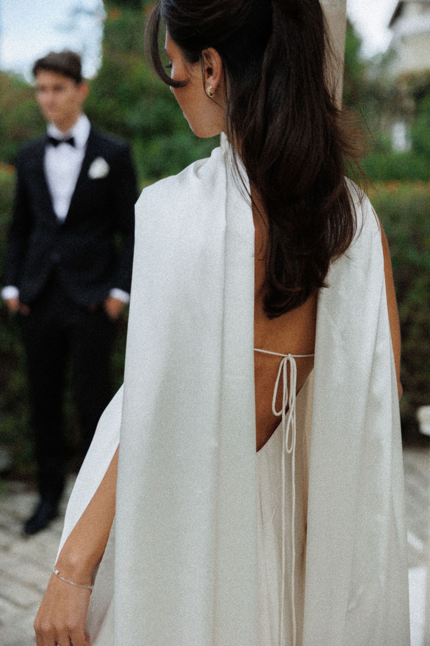 Elegant backless wedding dress with white cape and tie detail shown from behind with groom in tuxedo in background.