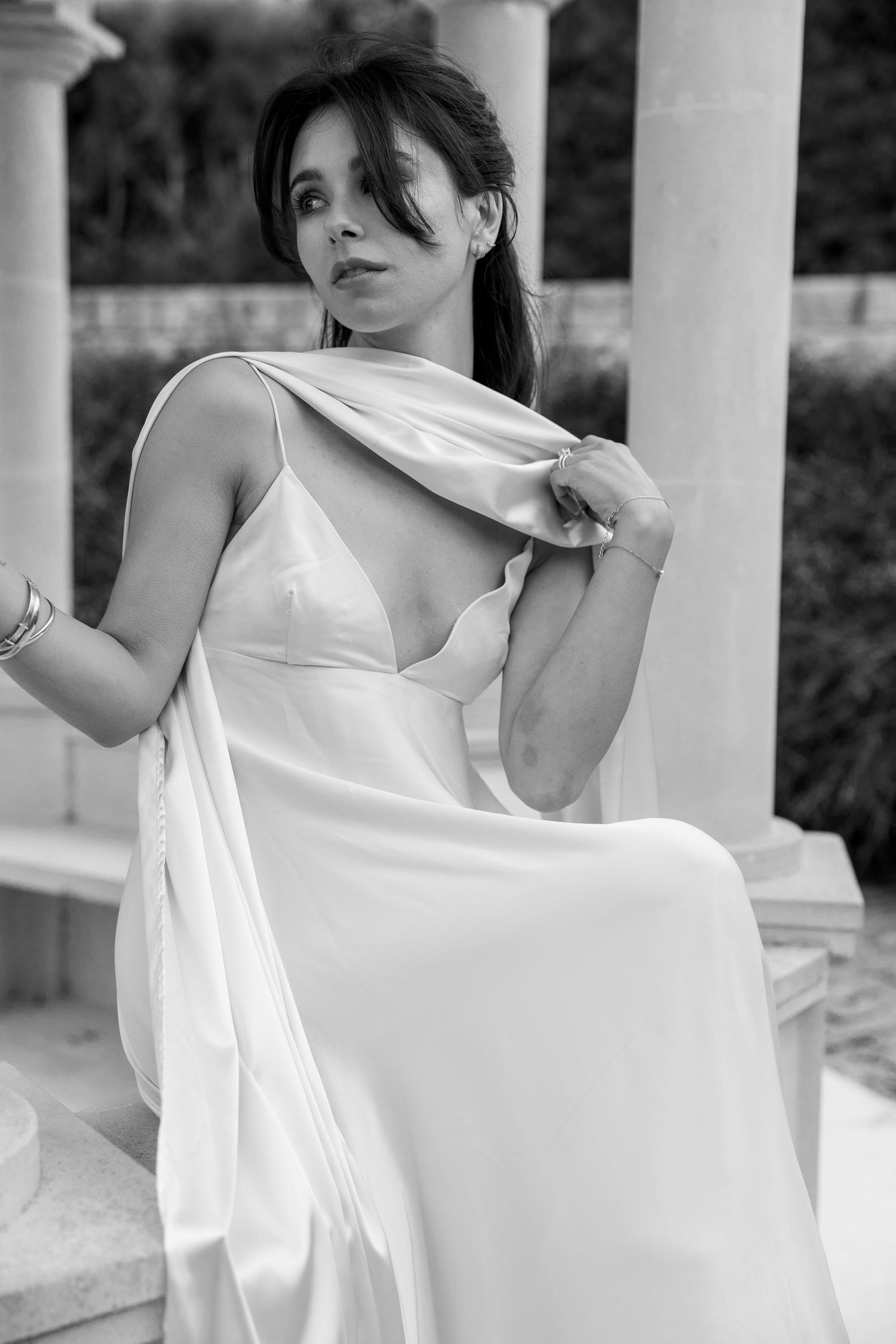 Black and white portrait of a woman in a flowing white dress posing elegantly by a pillar outdoors.