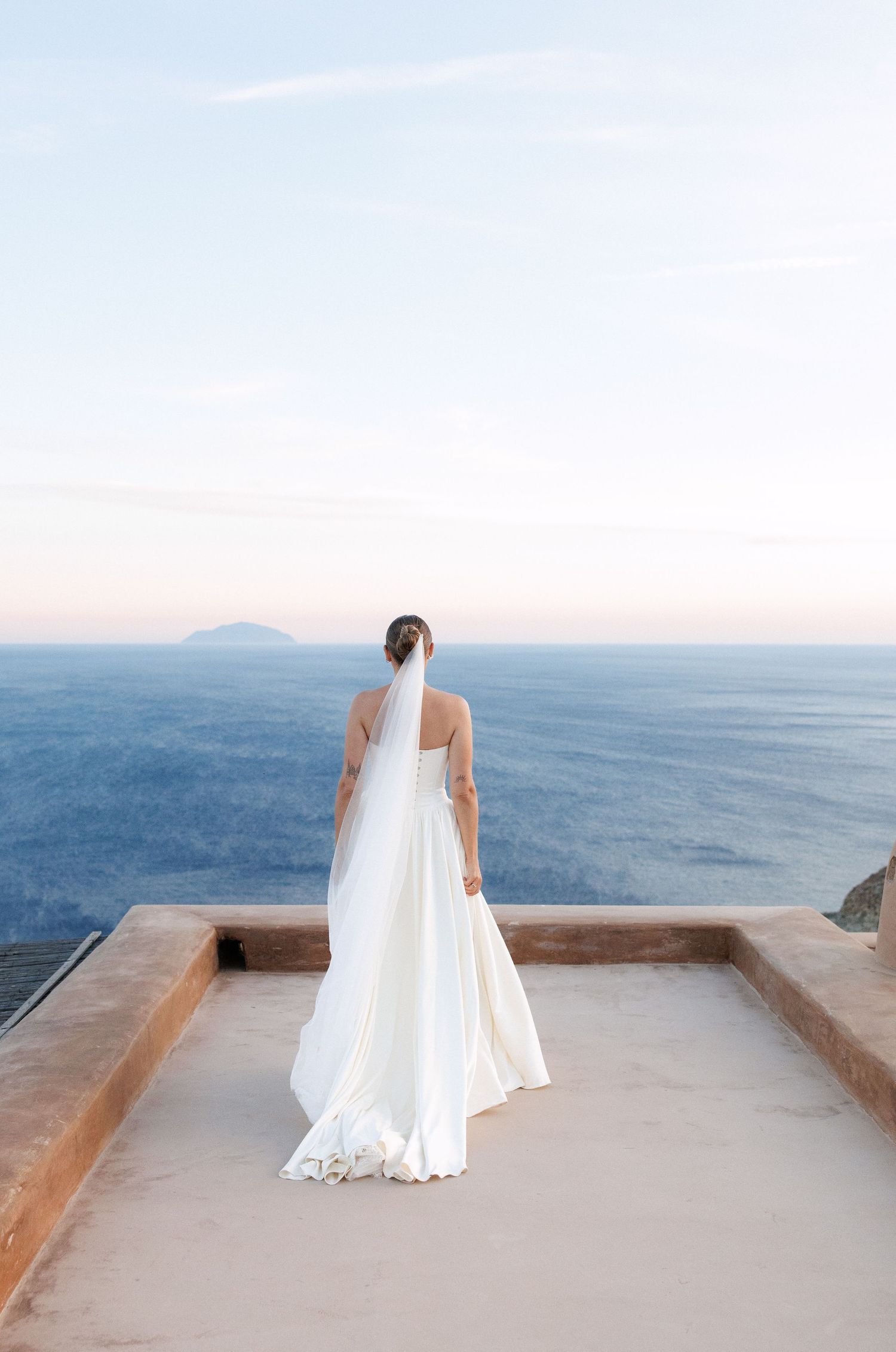 Bride in white gown and veil standing on terrace overlooking vast blue ocean at sunset.