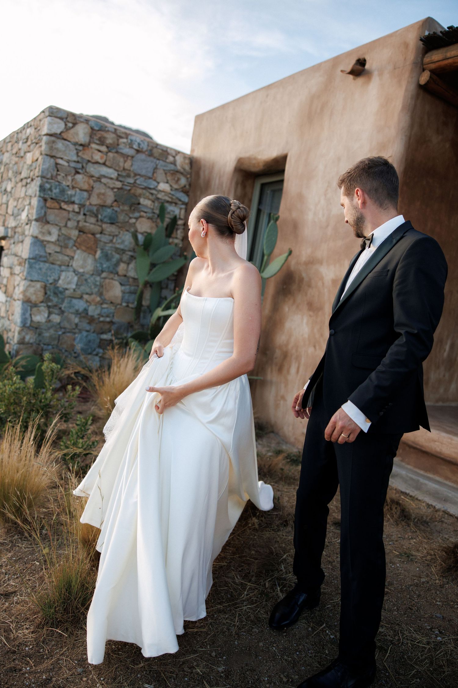 Bride in white strapless gown and groom in black tuxedo walk together near rustic stone building at outdoor wedding.