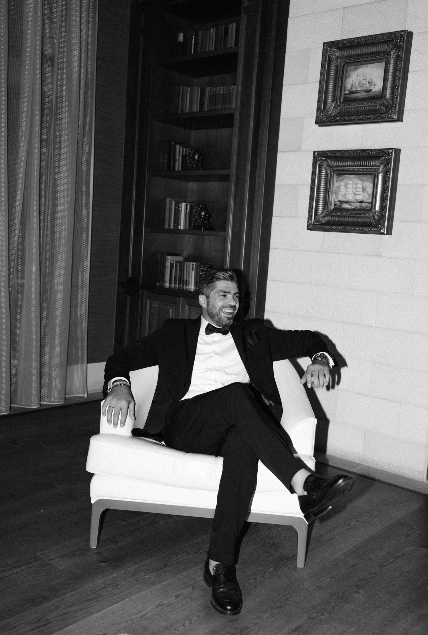 Man in tuxedo relaxing in white armchair, black and white photo with bookshelf and framed art.