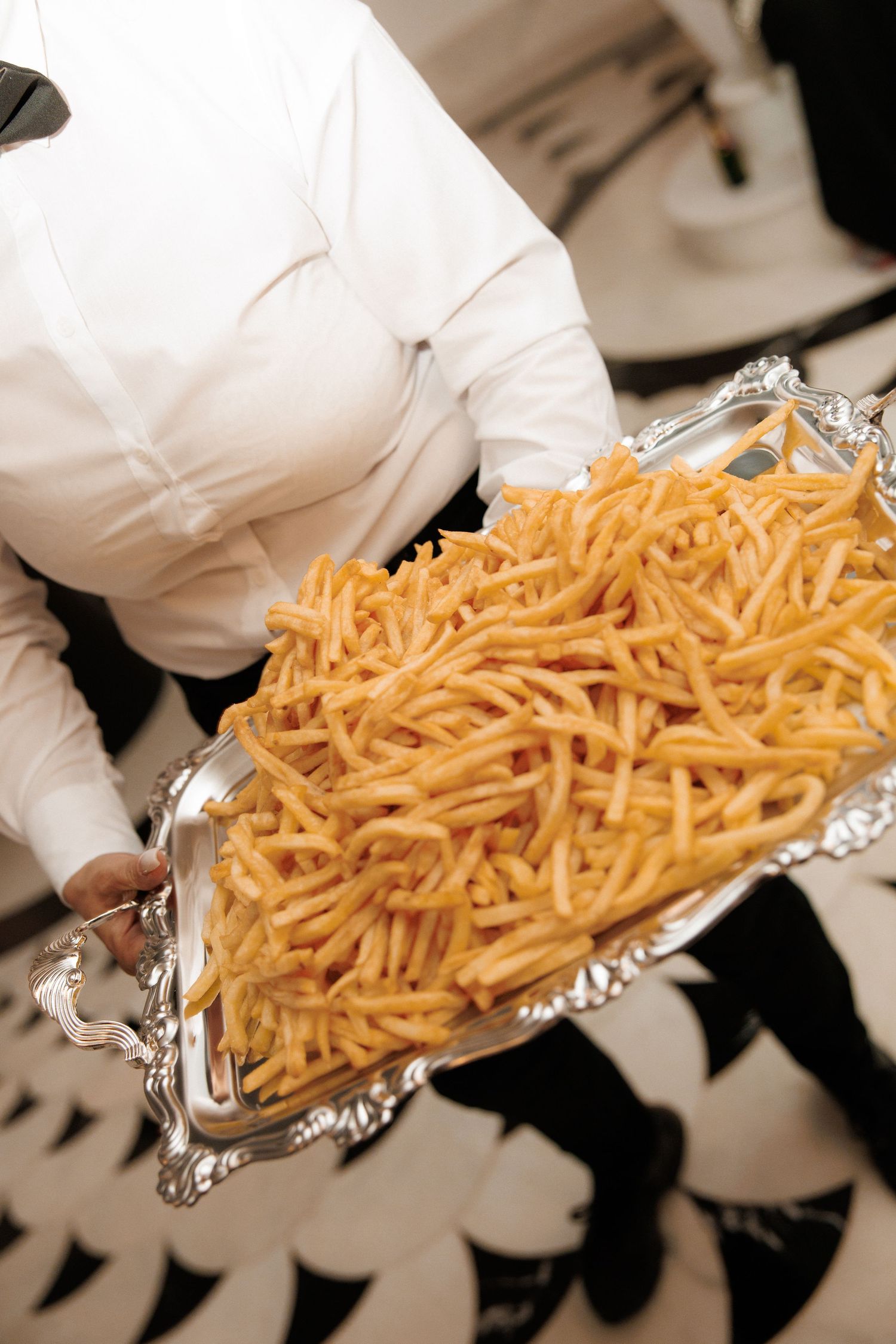 Waiter in white uniform serving a large silver tray overflowing with crispy golden french fries.