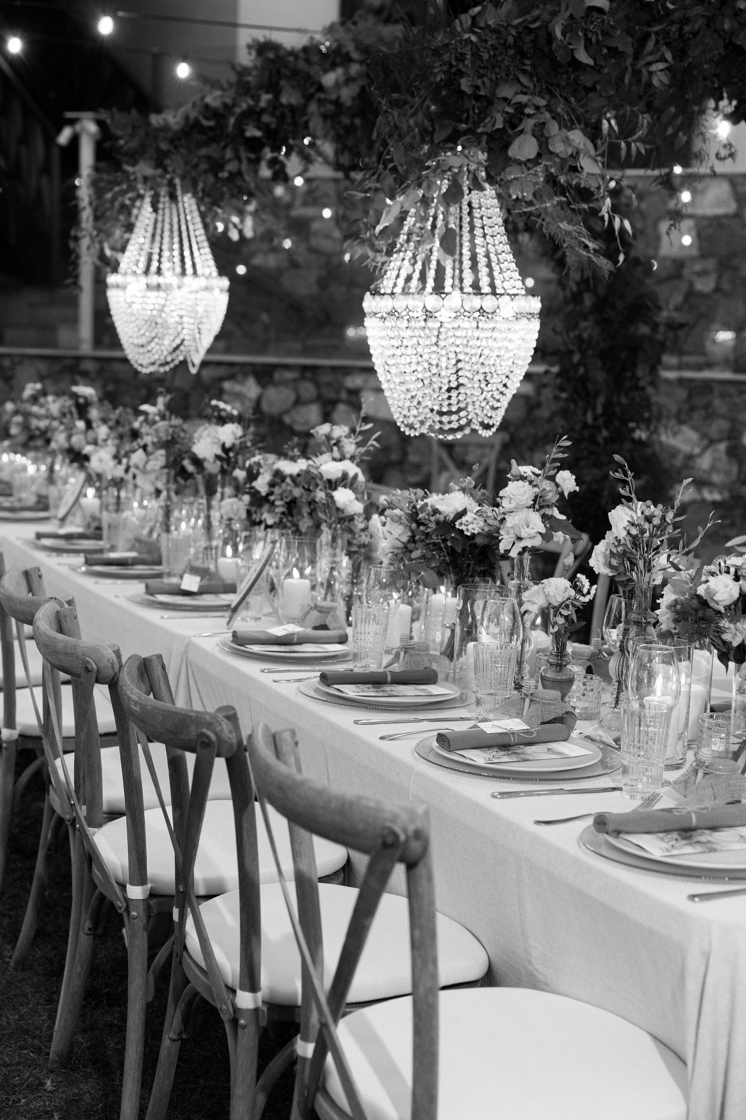 Black and white photo of elegant table setting with crystal chandeliers and cross-back chairs.