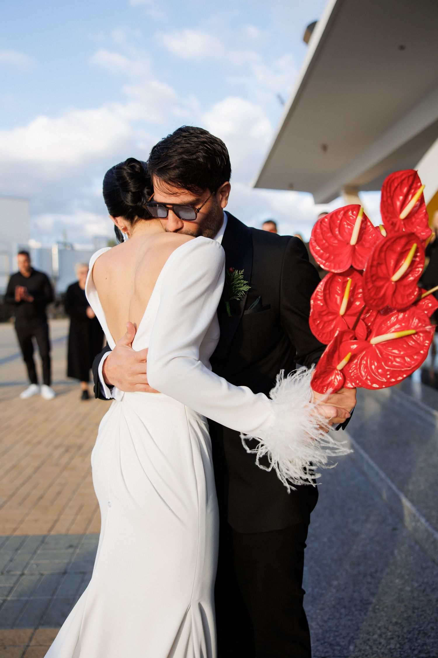 Bride and groom kissing outdoors, she holds red anthurium bouquet with feathers, wearing backless white gown and black tuxedo.