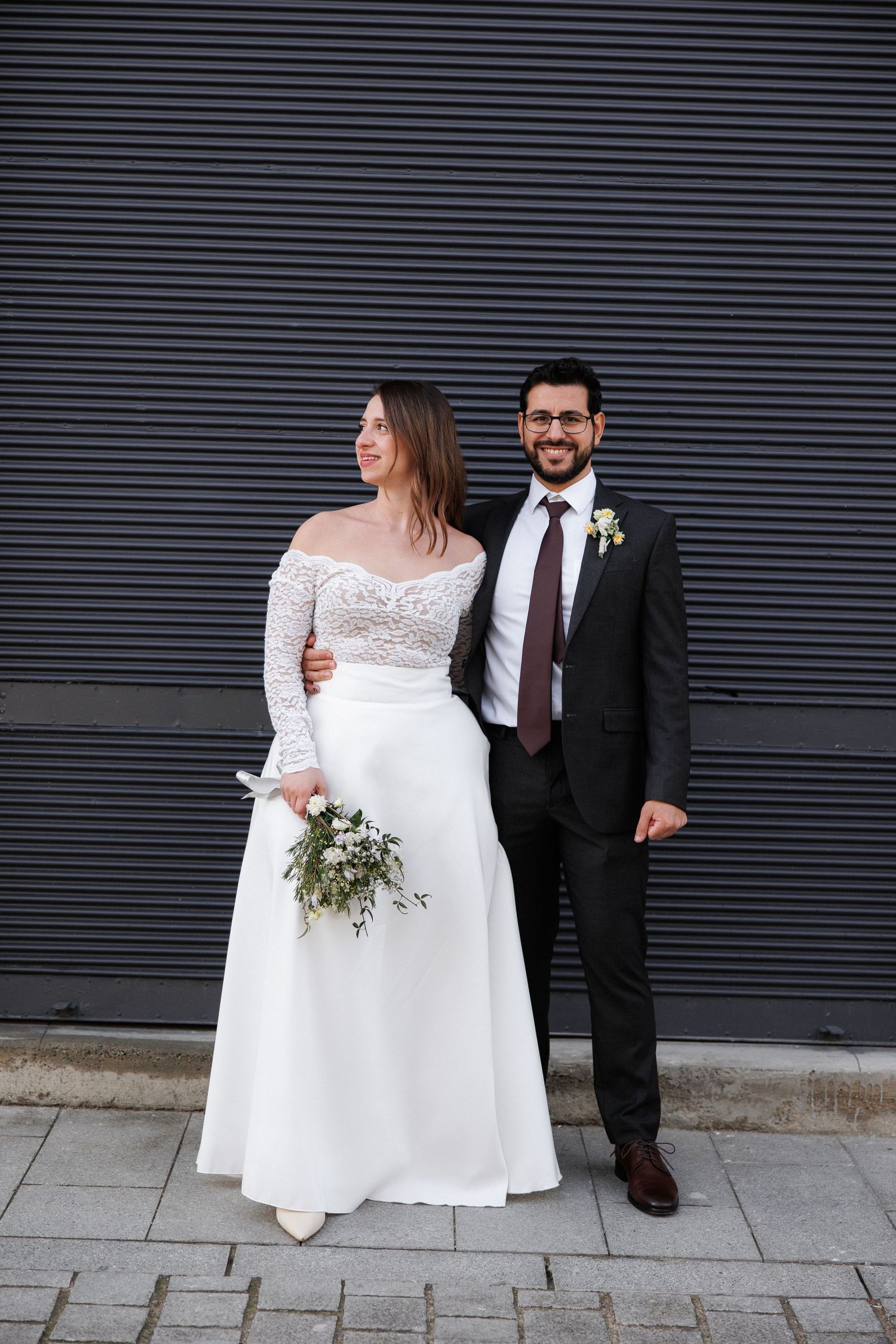 A bride in a white lace gown and groom in a dark suit with burgundy tie pose together outdoors on their wedding day.