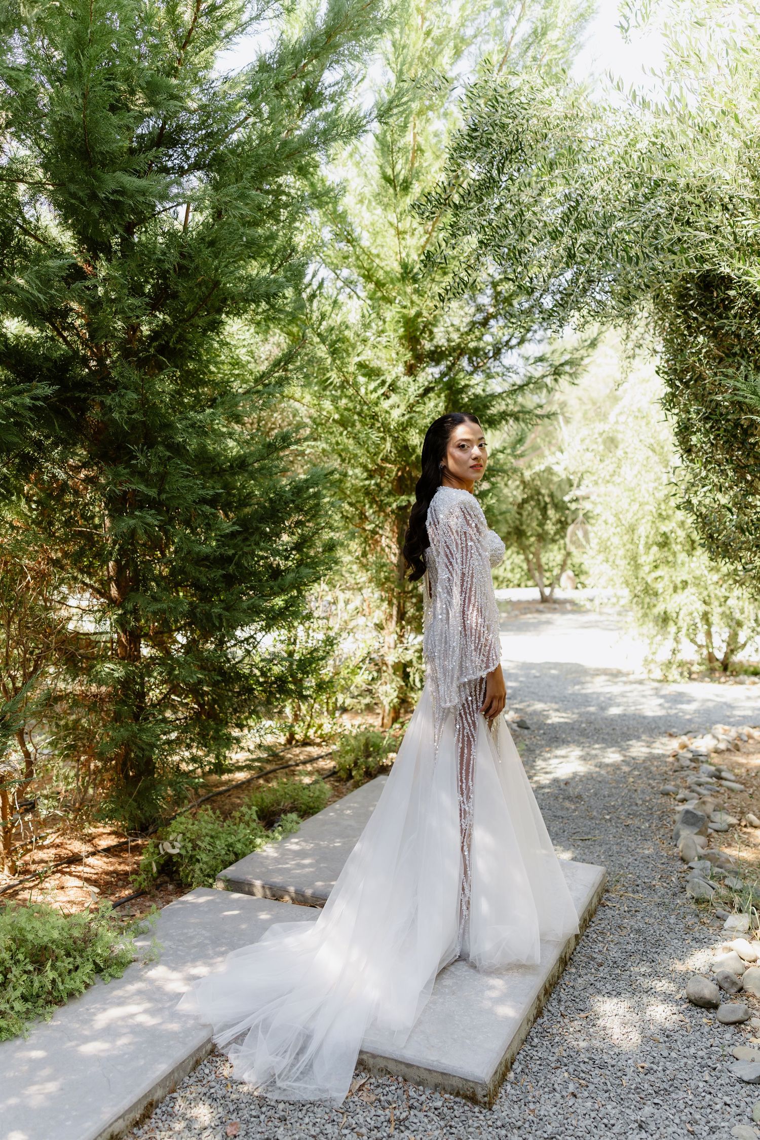 Bride in beaded long-sleeve wedding gown with flowing train stands on garden path surrounded by lush green trees.