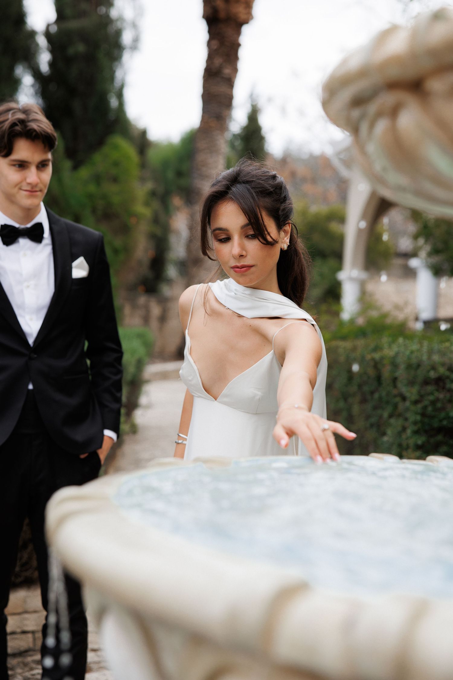 A candid wedding moment near a decorative fountain with cypress trees in the background.