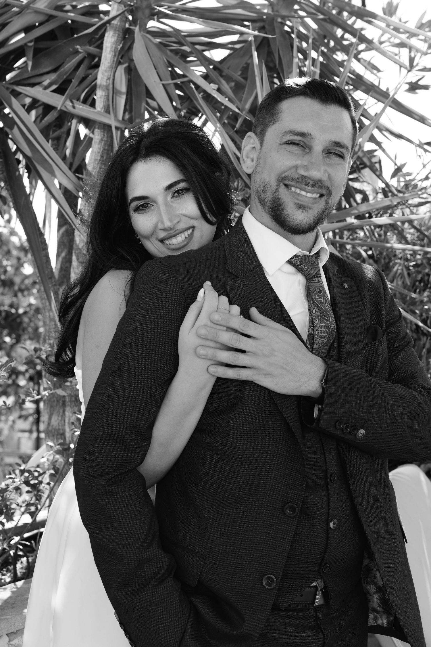 A romantic black and white portrait of a couple embracing outdoors with palm fronds in the background.