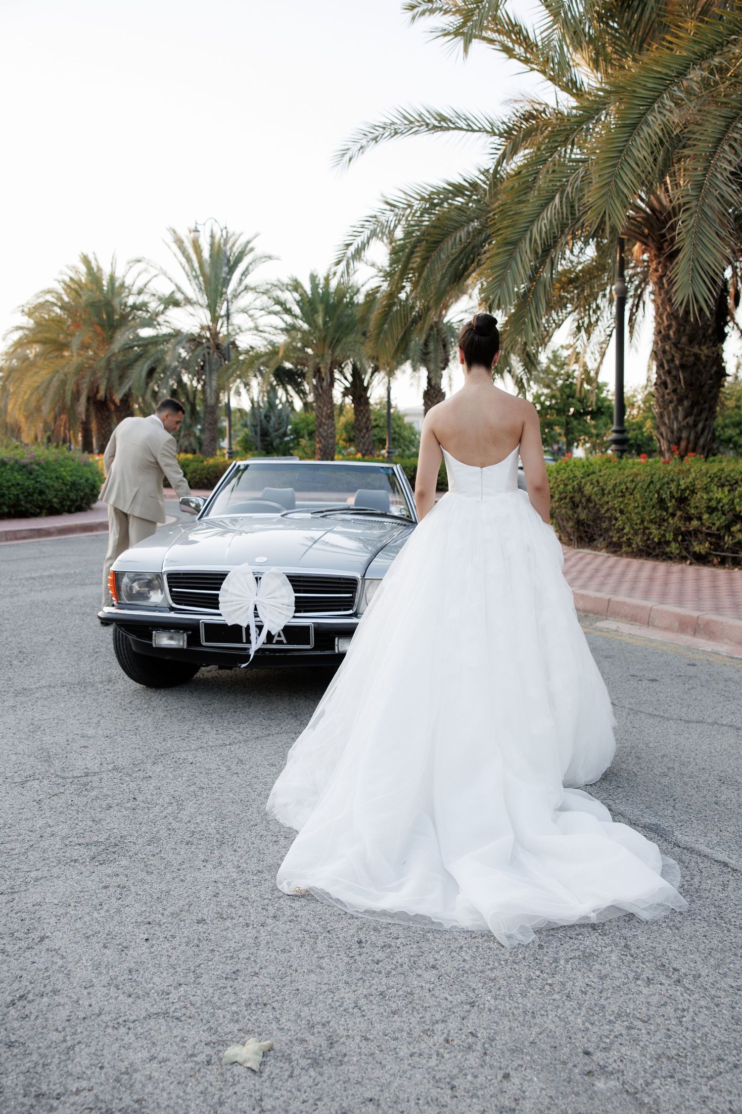 A bride in a white ballgown walking near palm trees and a parked vintage car.