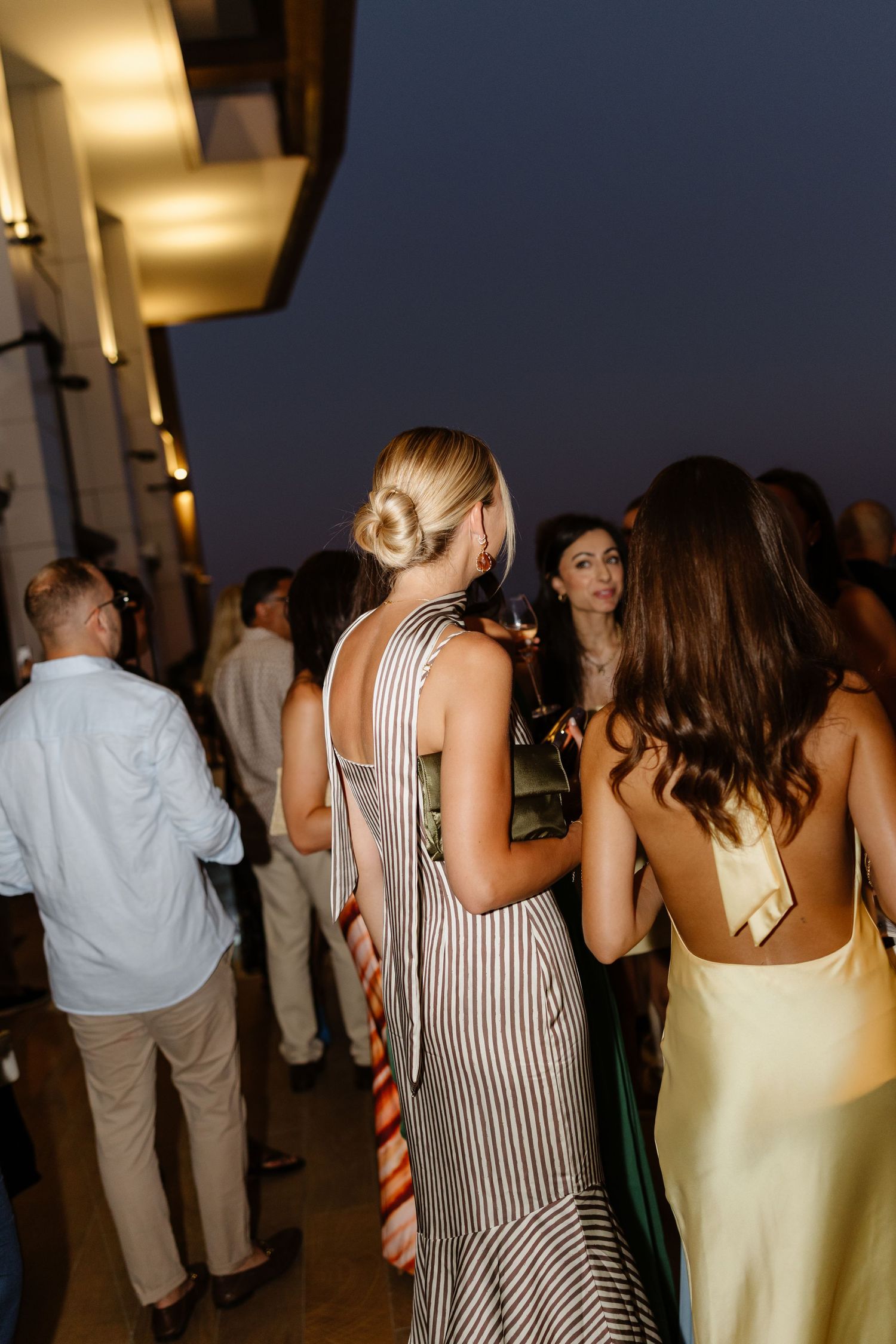 Guests in formal attire mingling at outdoor evening event with warm lighting.