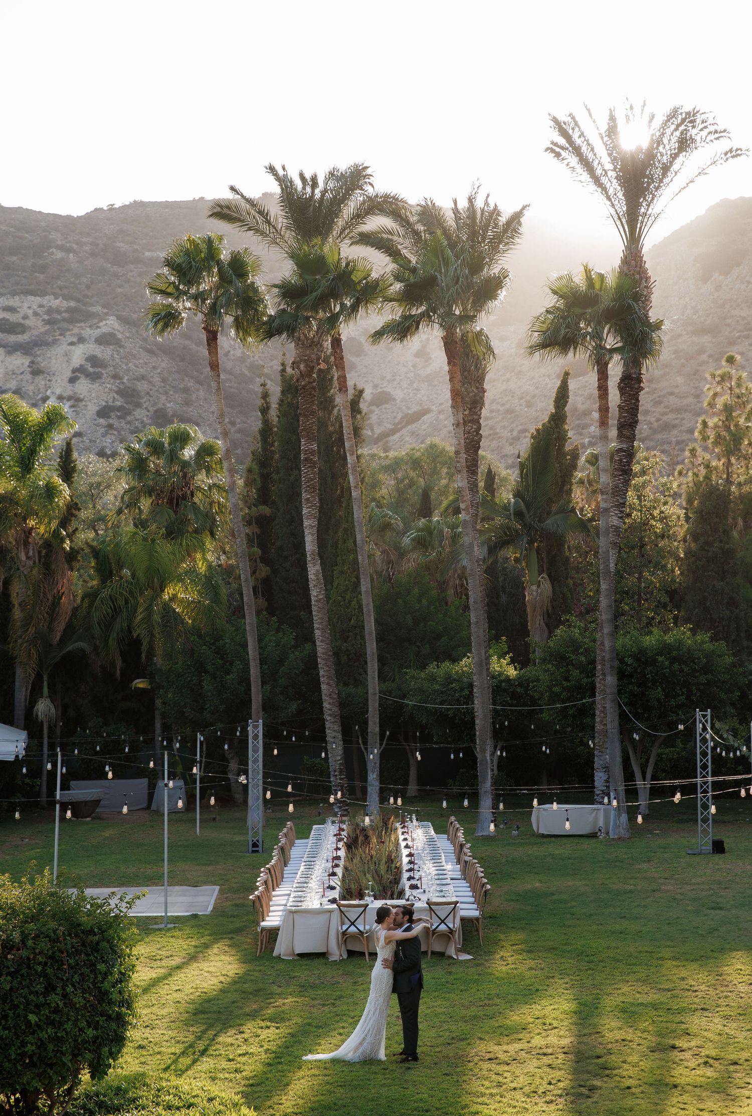 Couple sharing first dance at outdoor wedding reception with palm trees and mountain backdrop.
