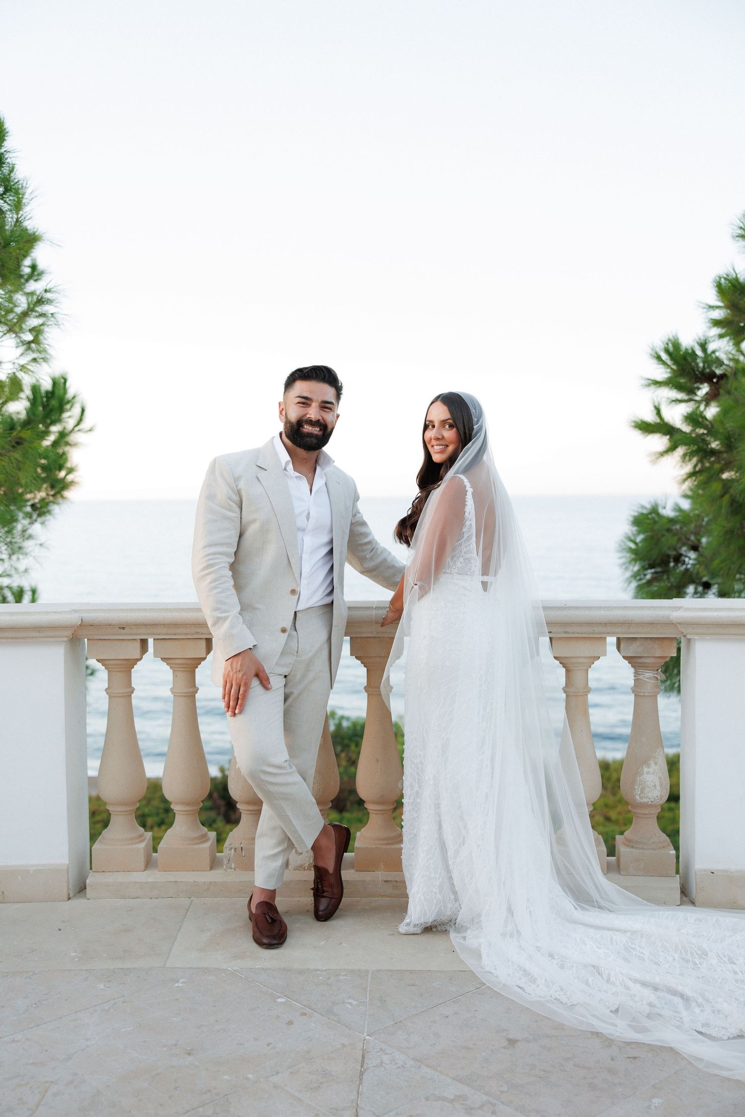Bride and groom pose on a balcony overlooking the ocean, bride wearing a long veil and lace gown.