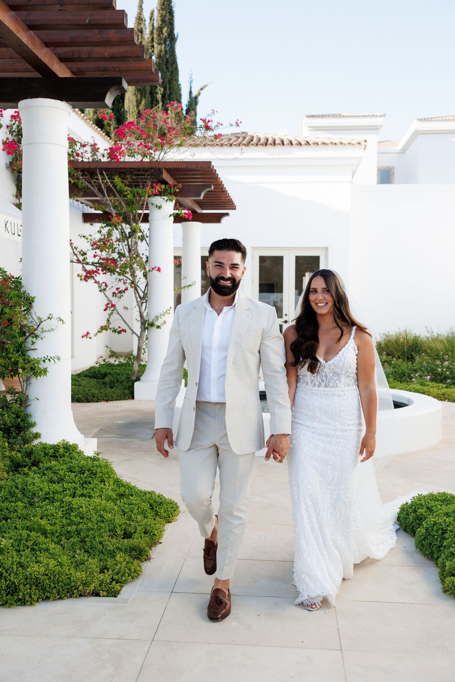 Bride and groom walking hand in hand at a white Mediterranean villa wedding venue with lush greenery.
