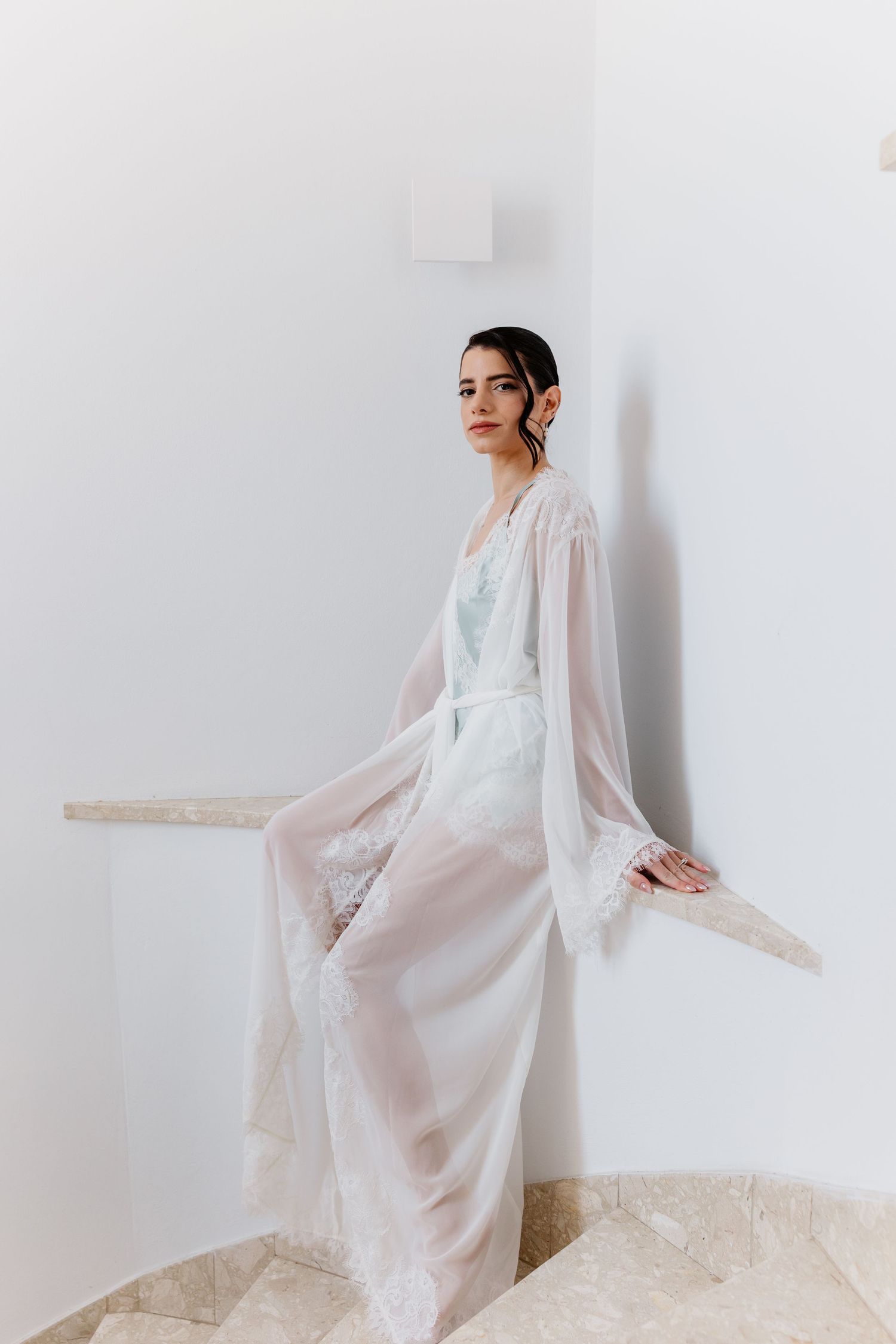 A bride wearing a sheer white lace robe over a light blue slip dress poses gracefully in a minimalist white room.