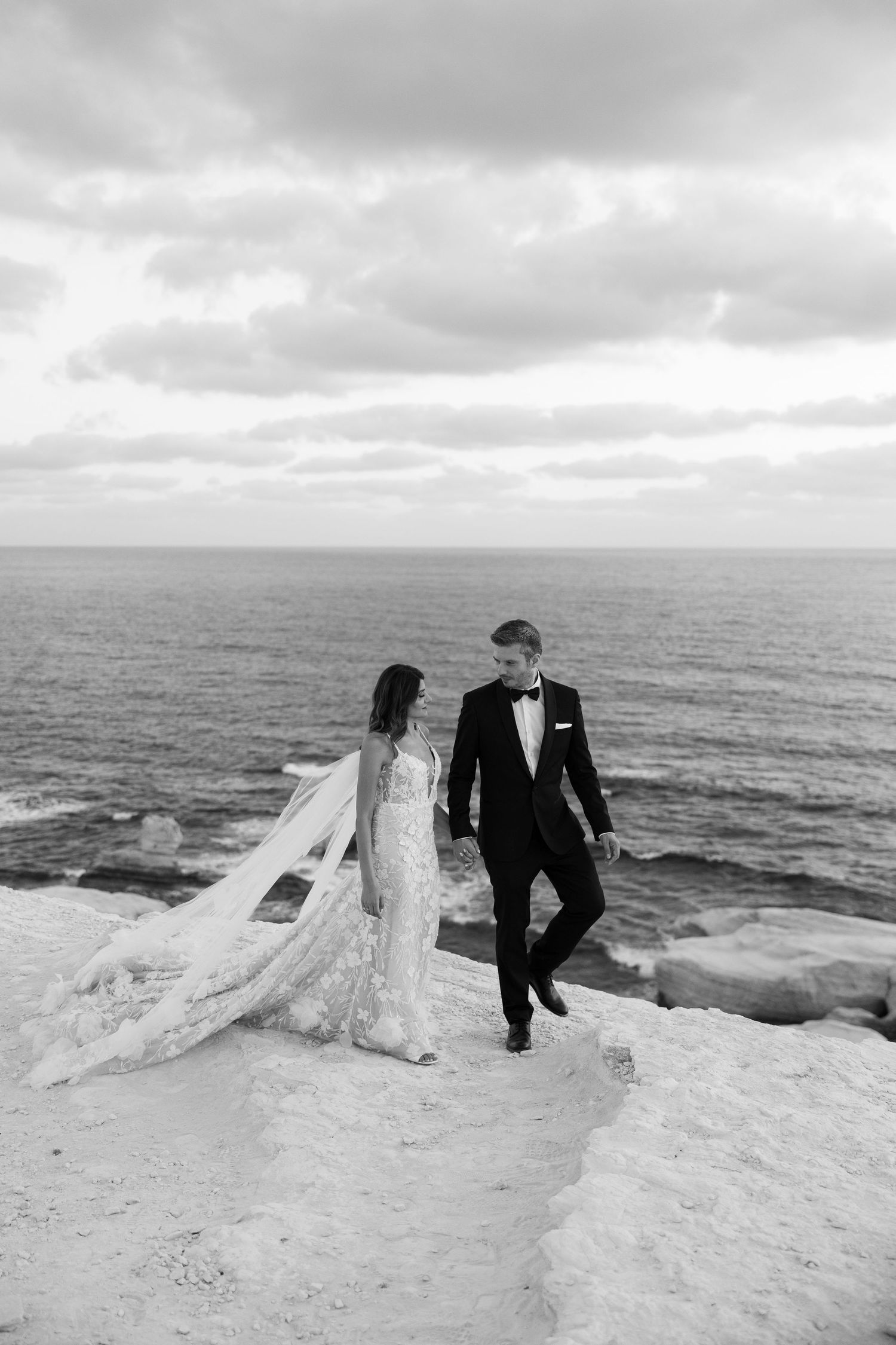 Black and white photo of a bride and groom holding hands on a rocky cliff overlooking the ocean on their wedding day.