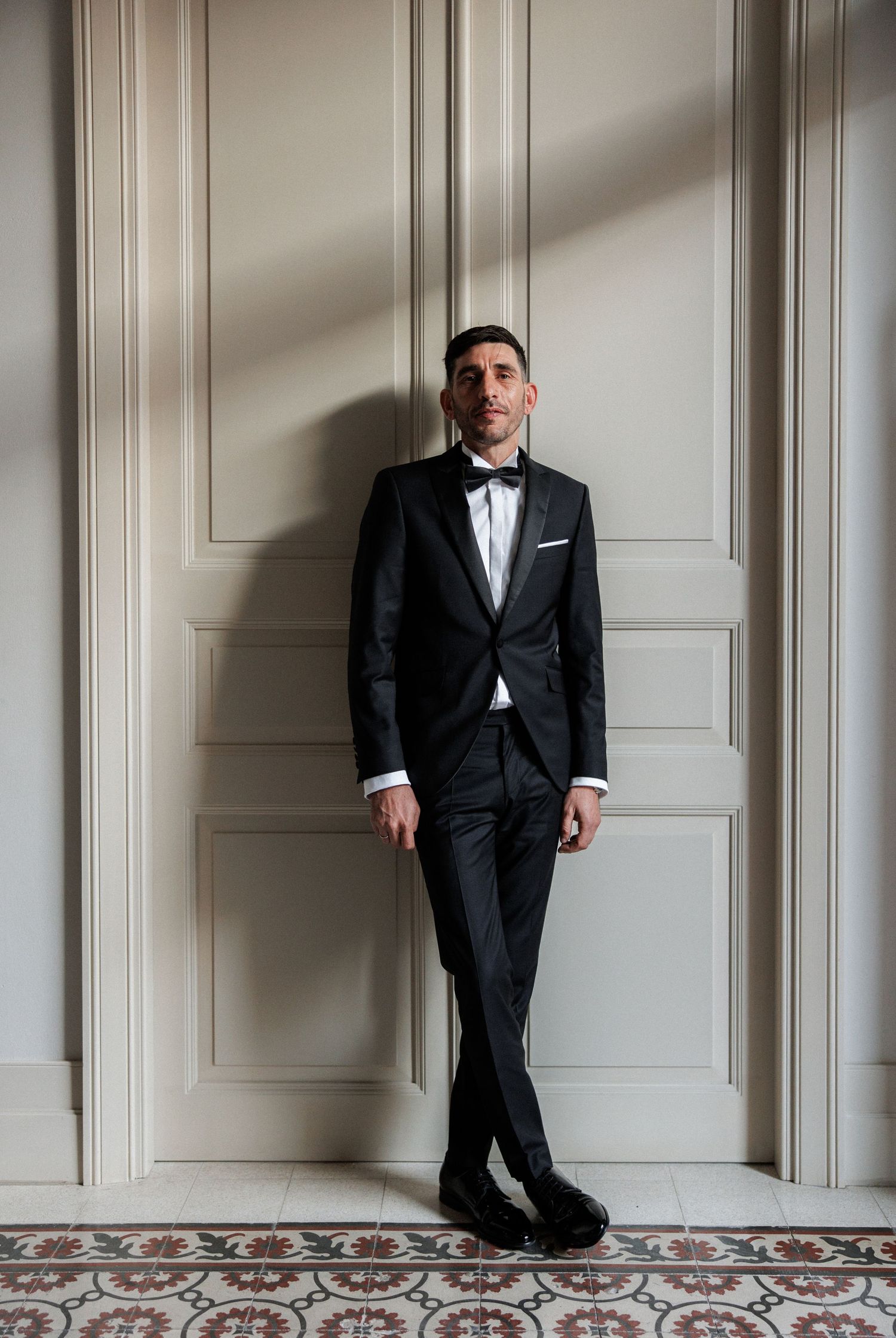 Man in black tuxedo with bow tie leaning against white paneled wall on decorative tile floor.