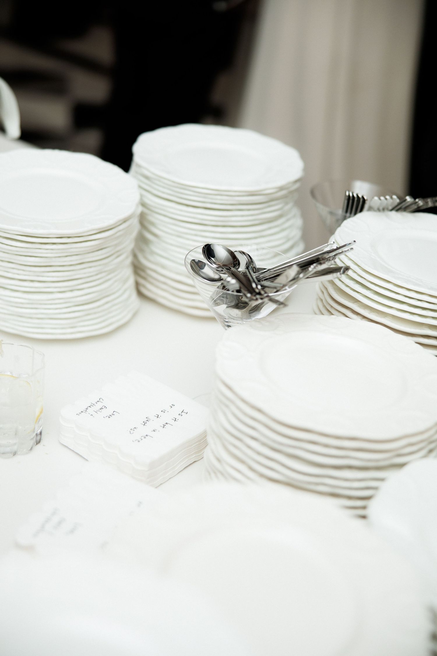 Stacks of white plates and silverware arranged on a white tablecloth for a catered event or wedding reception.