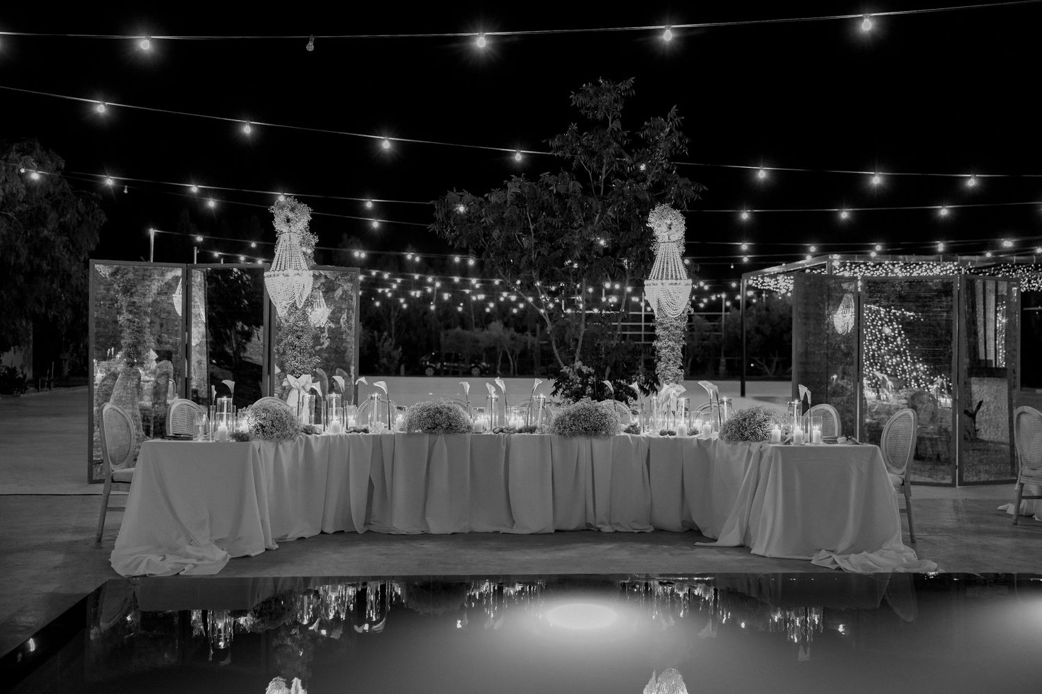 Elegant outdoor wedding reception setup with string lights and white tablecloths at night.