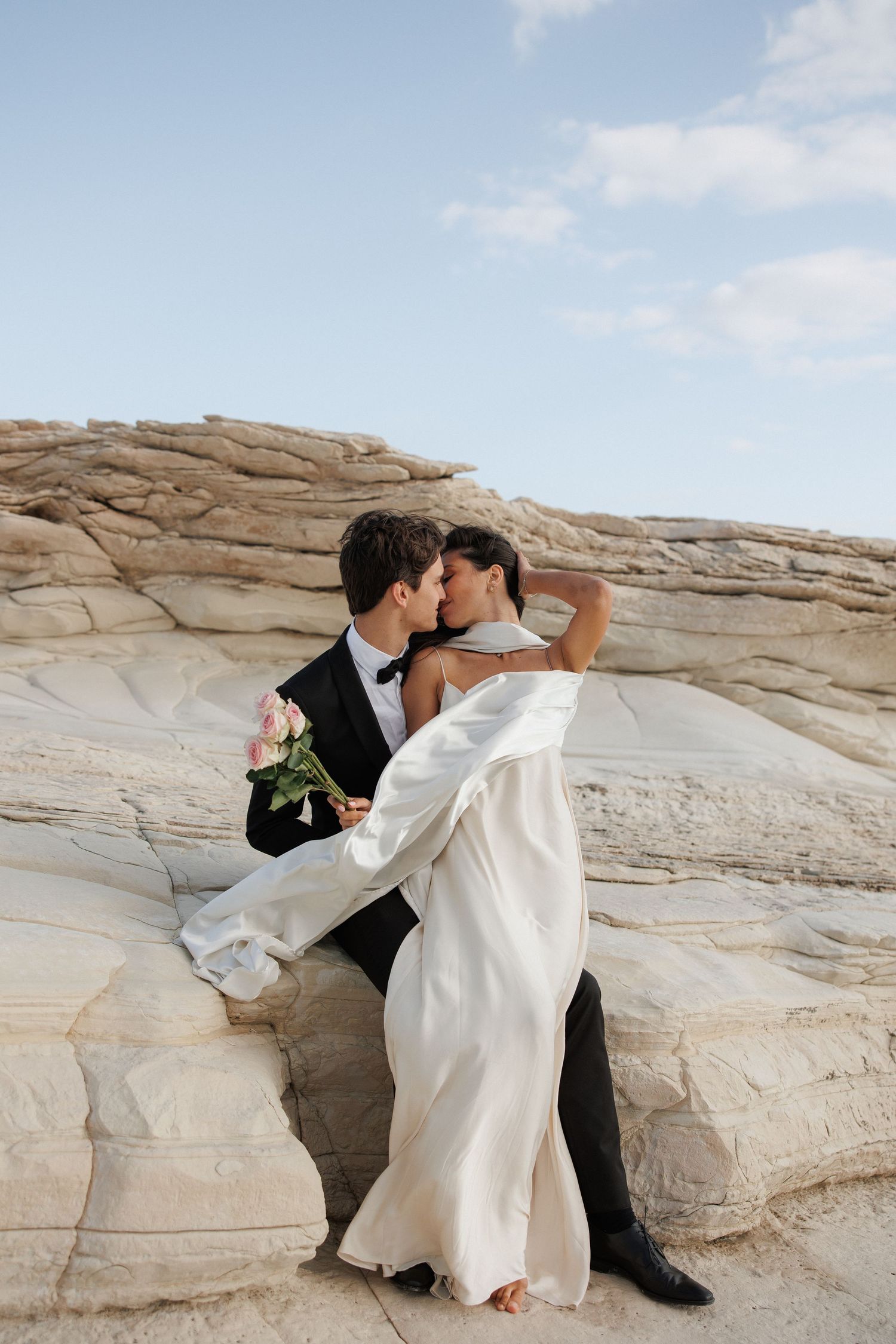 Couple in formal wedding attire embrace while standing on textured white rock formations under blue sky.
