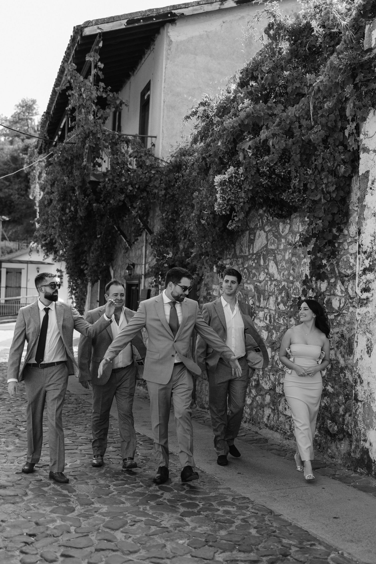 A group of people walking along a scenic cobblestone path beside an ivy-covered building wall in a vintage black and white photo.