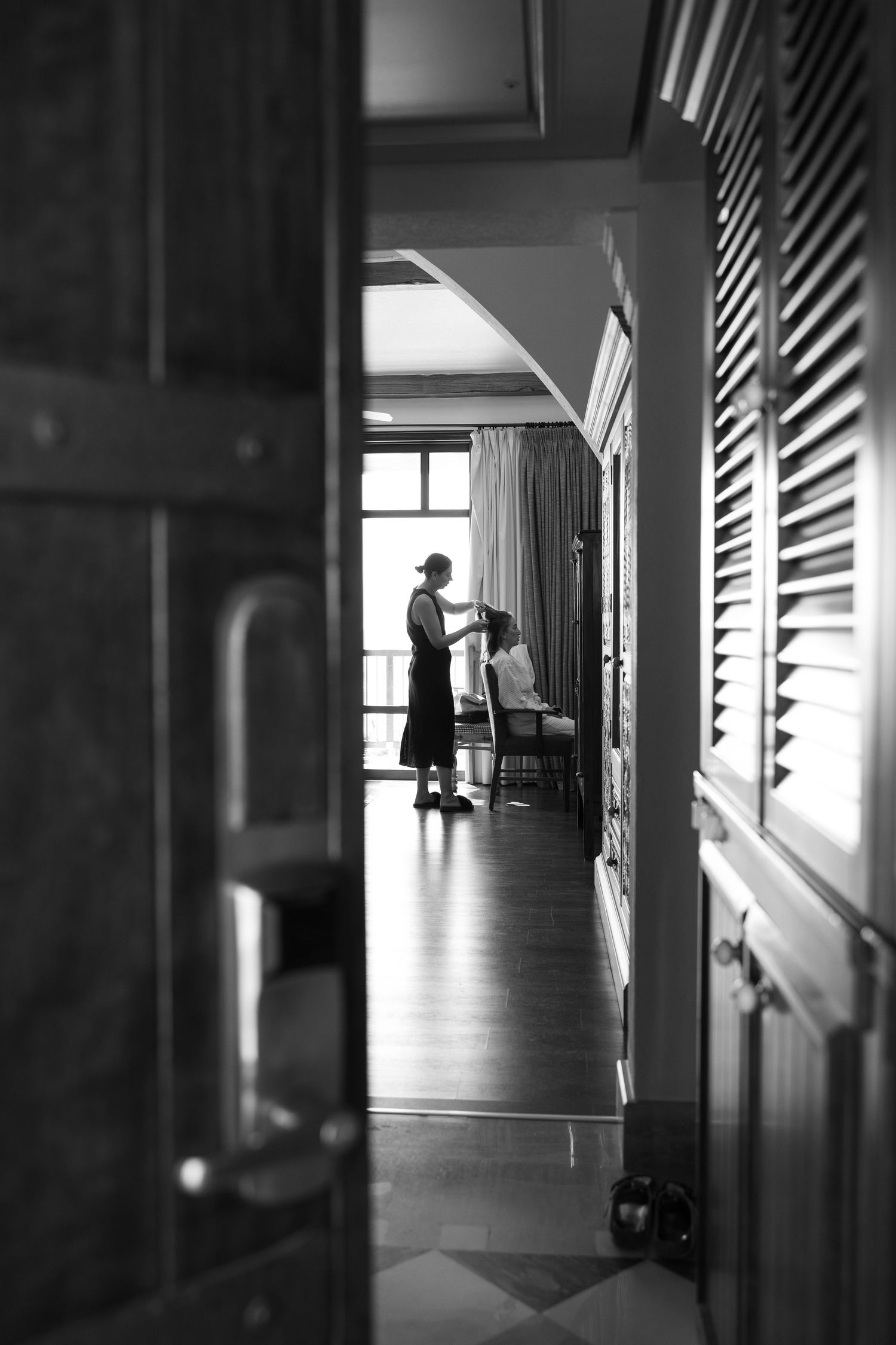 Black and white photo of a hairdresser styling a seated brides hair in an elegant hotel room before the wedding.