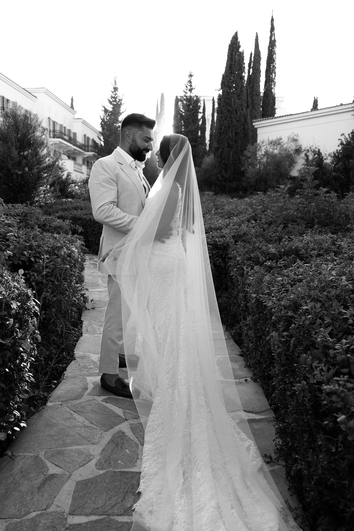 Black and white photo of bride and groom embracing in a garden pathway, bride wearing a long veil and lace gown.