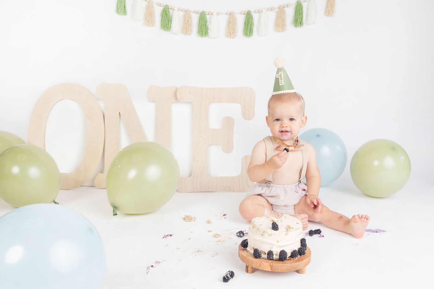 Baby in party hat smashes first birthday cake surrounded by green and blue balloons and ONE sign.