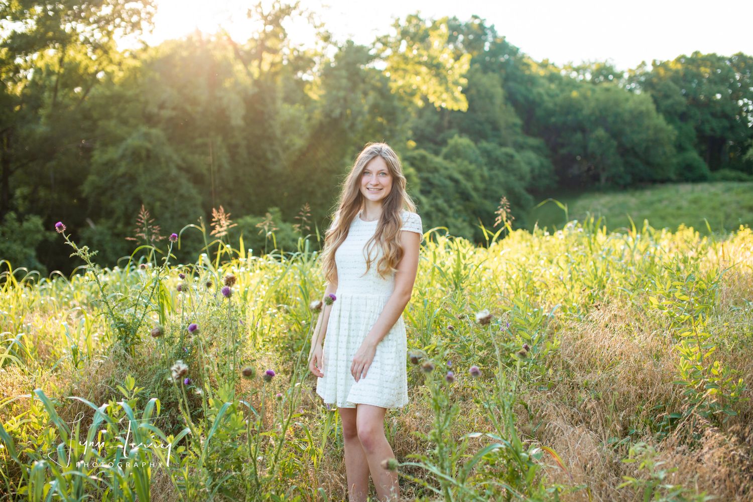 Young woman in white dress standing in a sunlit wildflower meadow with lush green trees in the background.