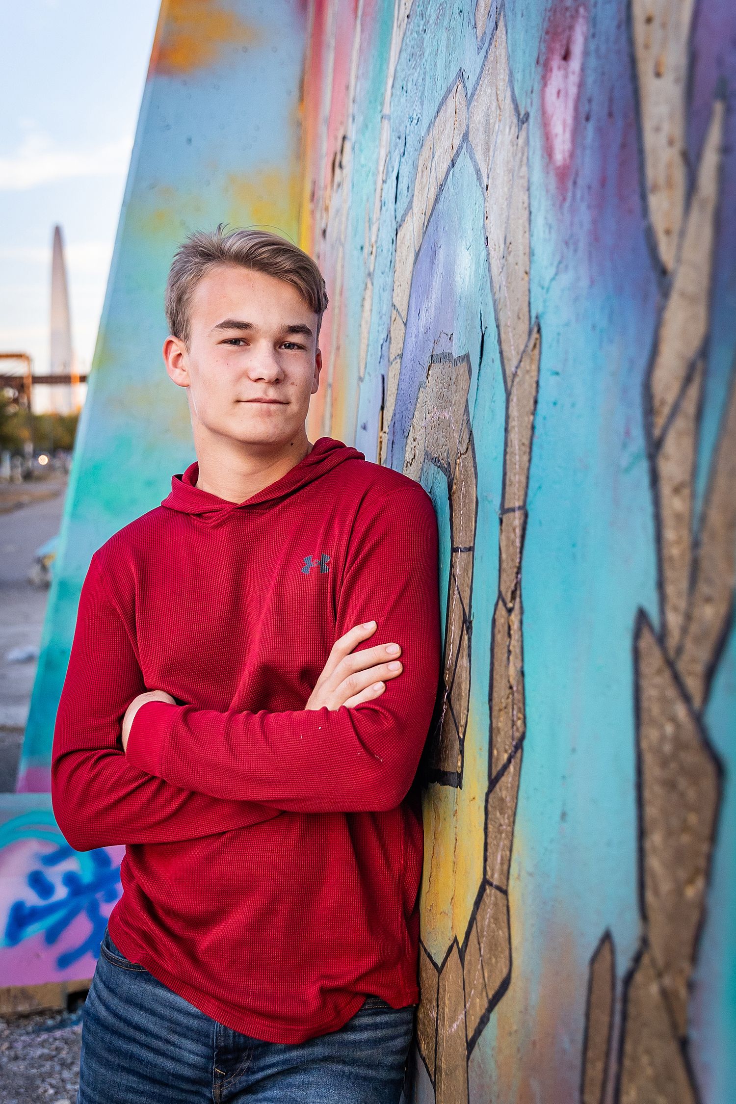 A teenage boy in a red hoodie with crossed arms leans against a colorful graffiti mural outdoors.