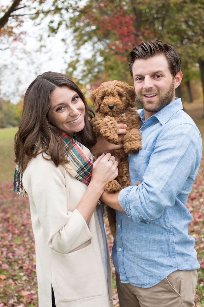 A couple in fall attire holds their brown goldendoodle puppy while standing among autumn leaves.