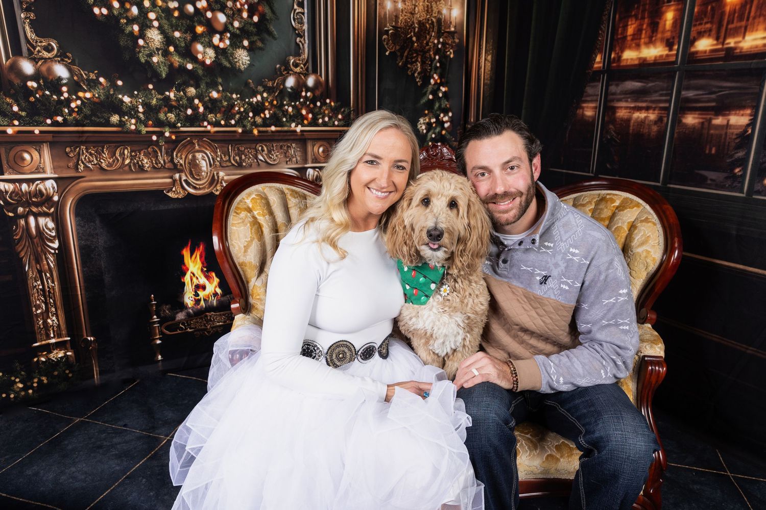 Couple in festive attire with golden doodle dog wearing bow tie, sitting by decorated fireplace for Christmas portrait.