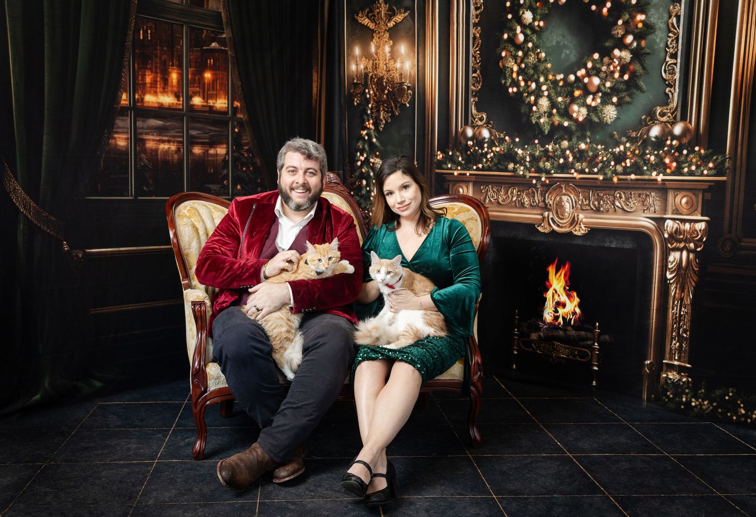 Couple sitting on red velvet chair by fireplace holding cats in festive Christmas decorated room.