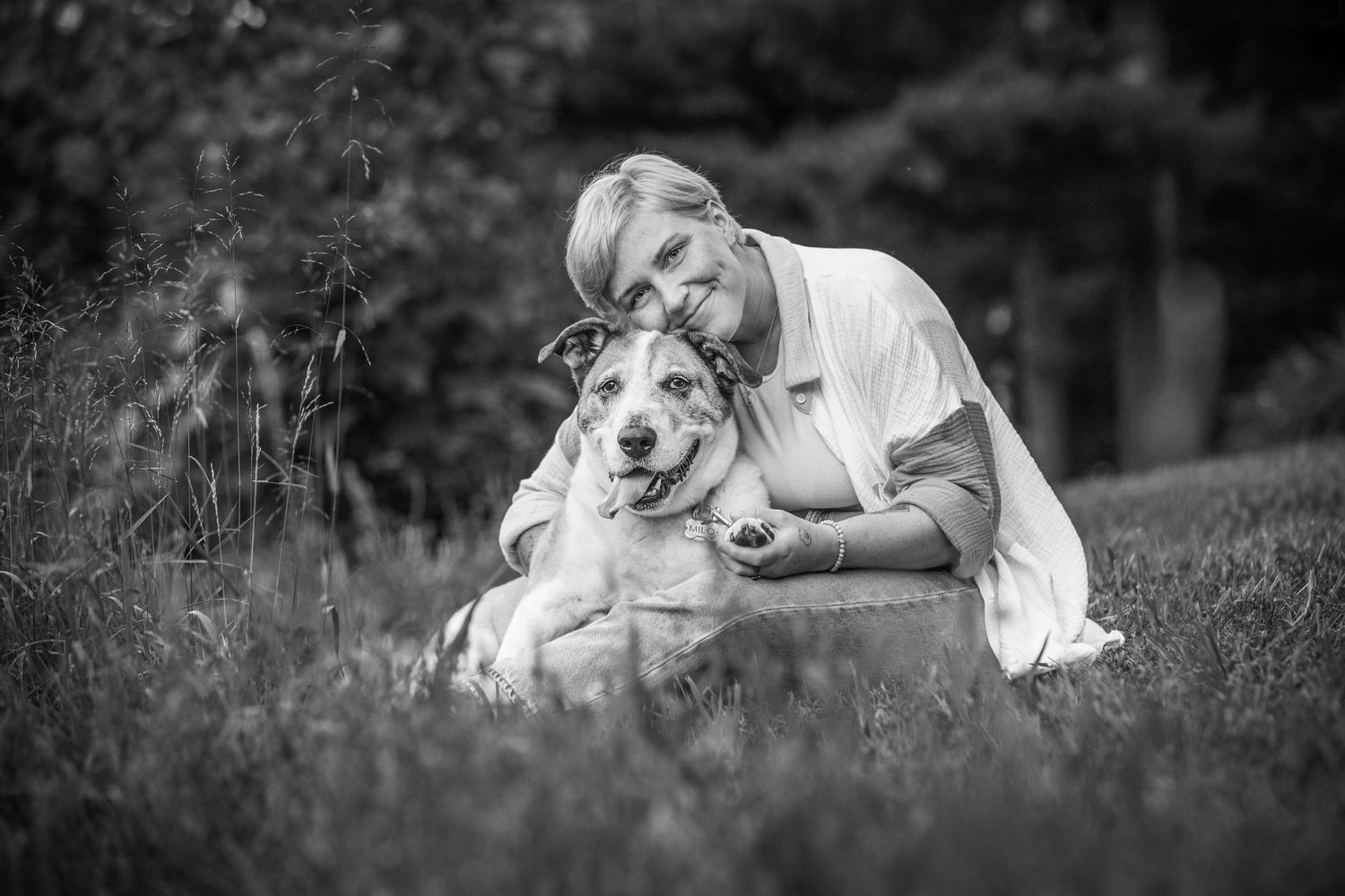 Black and white photo of a smiling woman hugging her large dog while sitting in a grassy park.