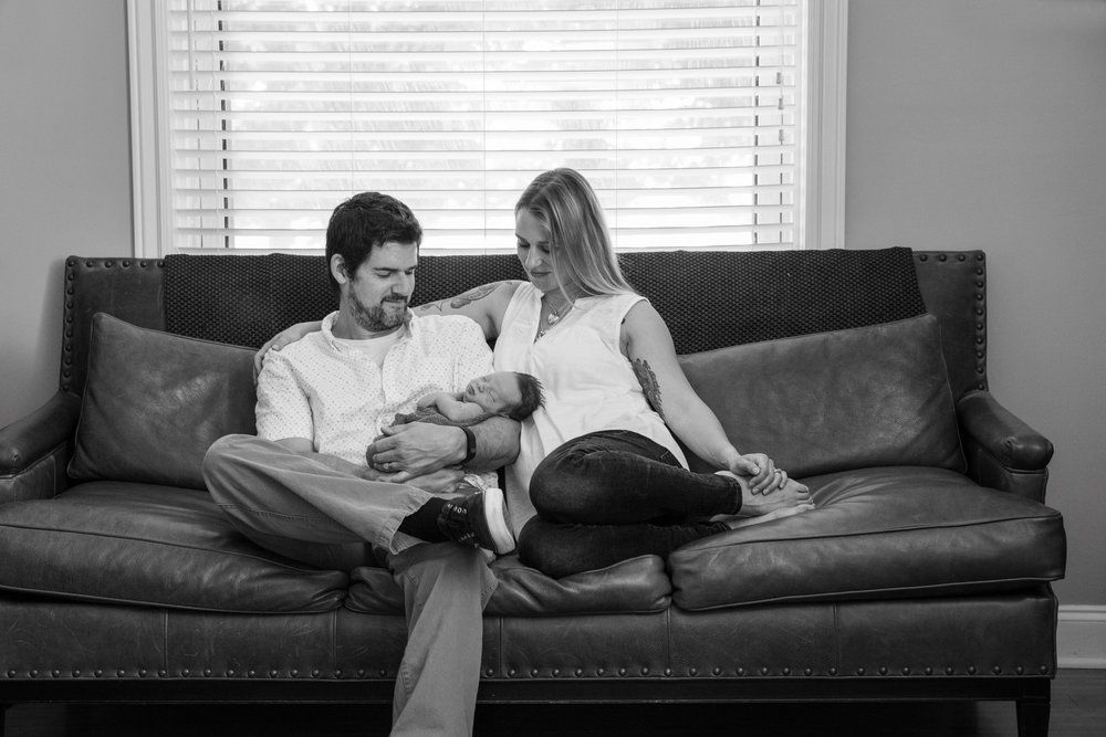 Couple sitting together on gray couch looking at newborn baby.