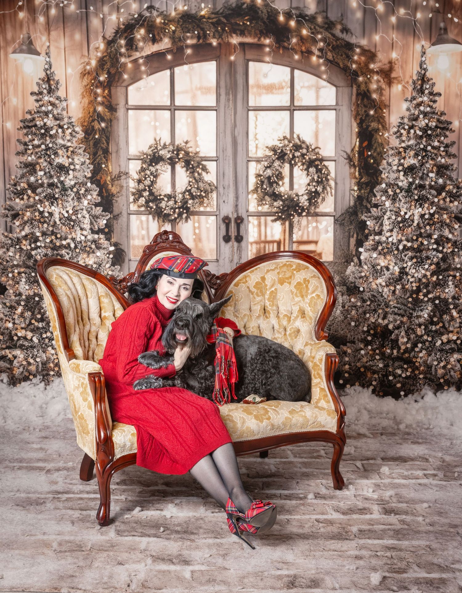 Woman in red dress with black dogs on ornate sofa, surrounded by Christmas trees and wreaths in festive holiday setting.