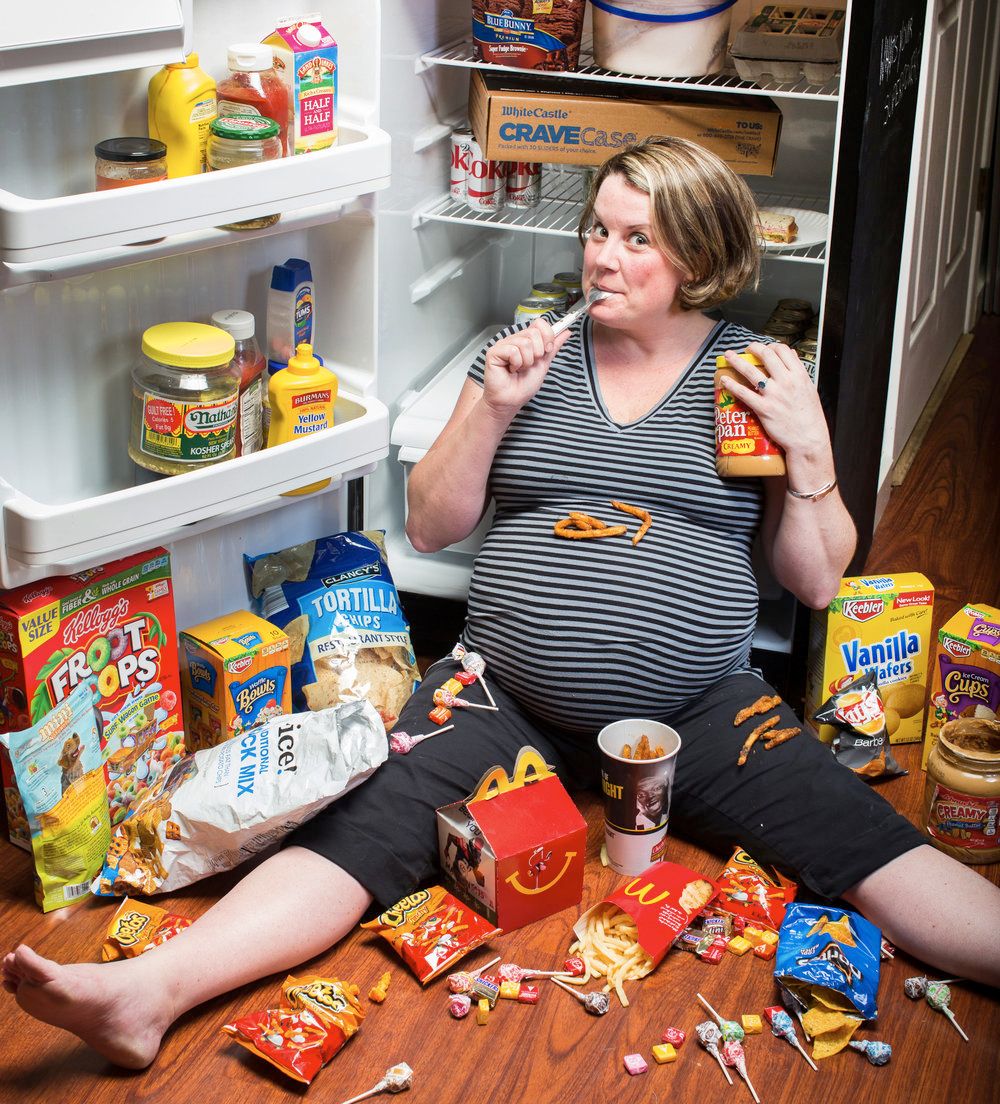 Person sitting on floor surrounded by junk food and snacks with open refrigerator in background.