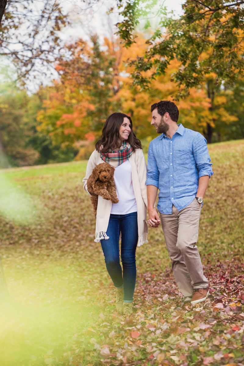 A couple walks through a park with autumn foliage holding their goldendoodle puppy.