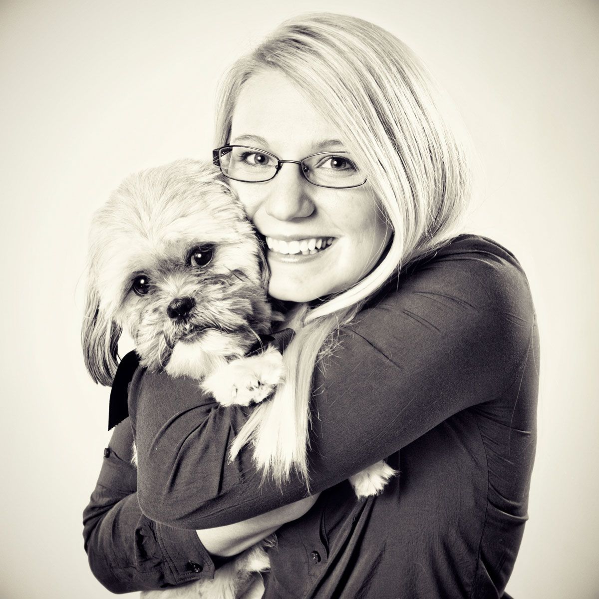Black and white portrait of someone holding a small fluffy white dog while wearing glasses.
